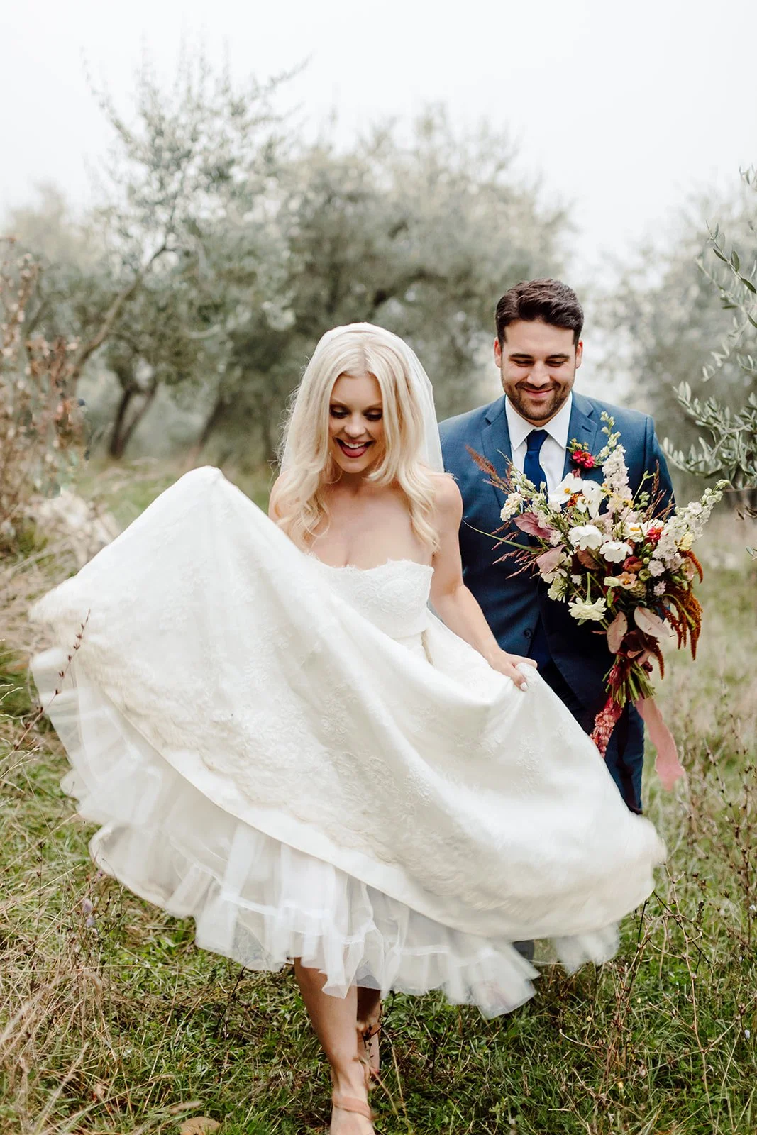 A bride and groom walking outdoors in a natural, grassy setting with trees in the background. The bride is wearing a white dress and holding it up while smiling, and the groom is in a blue suit holding a bouquet of flowers, also smiling.