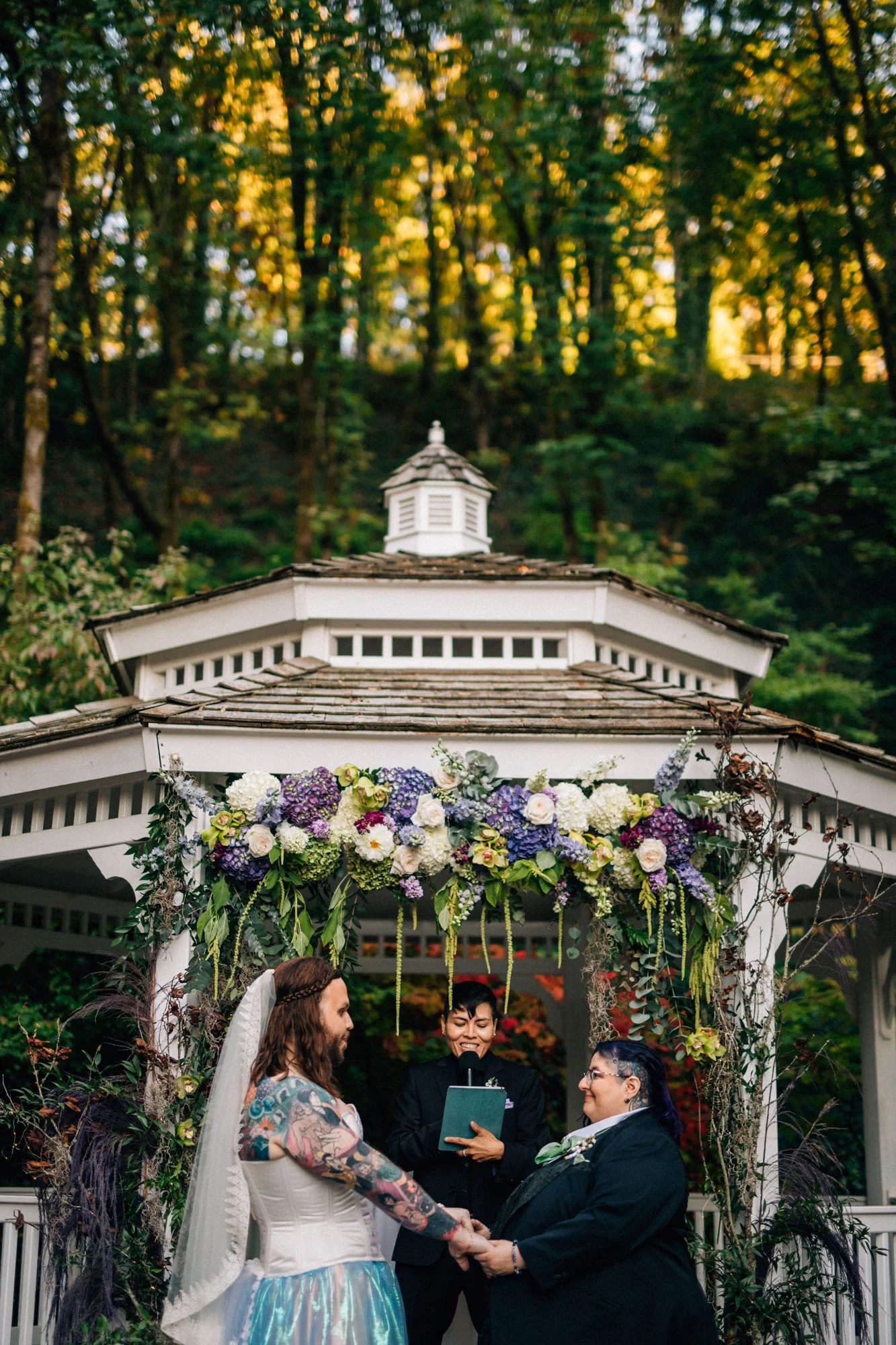 A couple and an officiant stand under a floral archway and gazebo in a forested setting. One member of the couple has long brown hair, tattoos, and is wearing a white and blue gown. The other member of the couple is wearing a black suit and glasses.