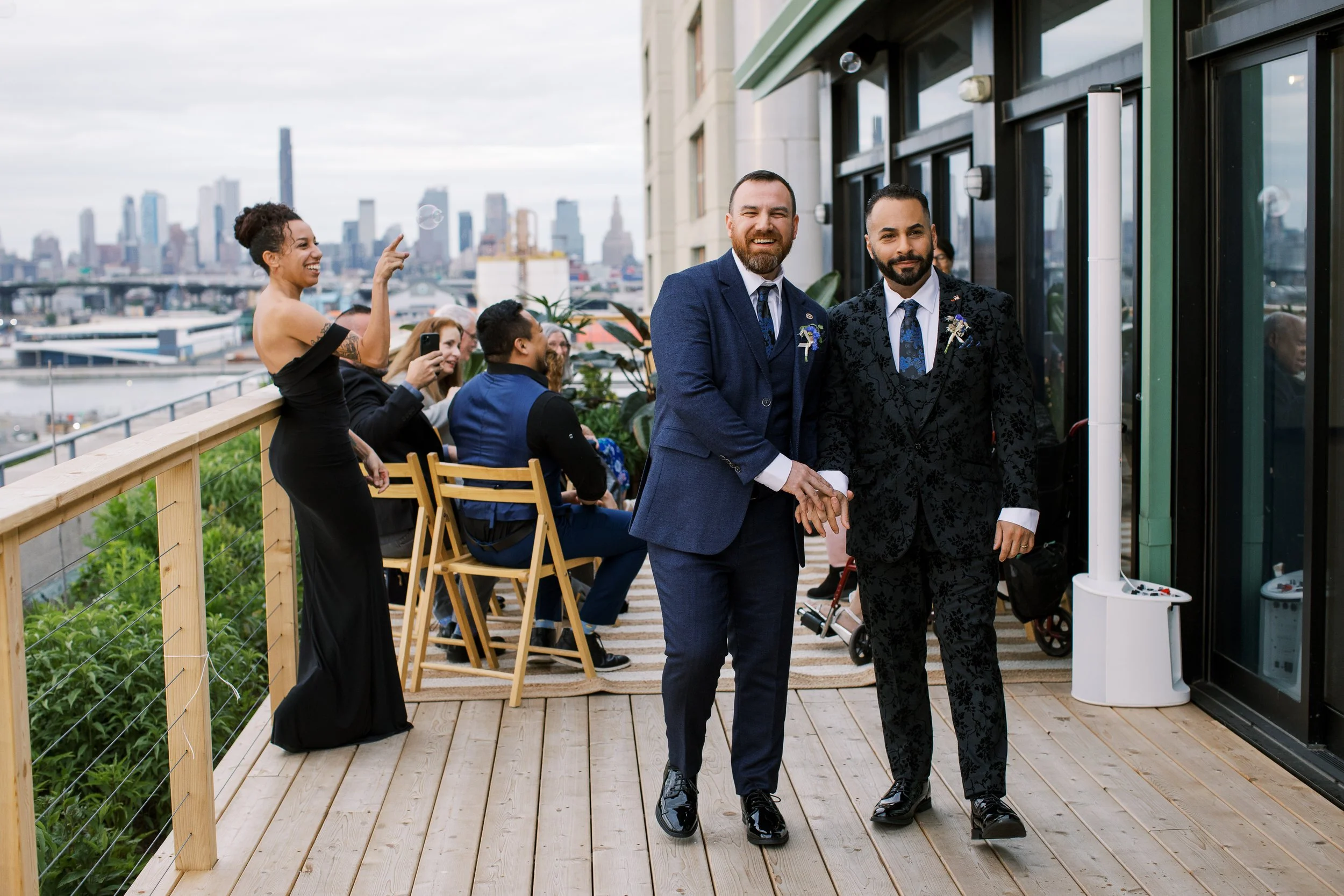 Two men in suits holding hands and smiling at a rooftop wedding ceremony, with guests sitting and standing nearby in an urban cityscape background.