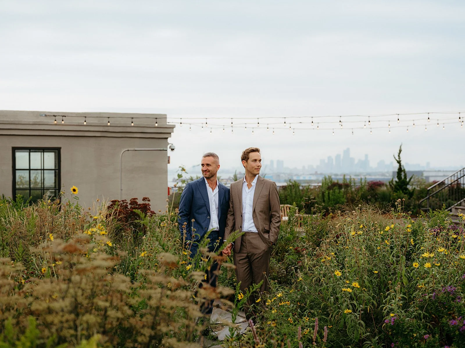 Two men standing on a rooftop surrounded by flowers. One wears a medium blue suit and white dress shirt. The other wears a brown suit and white dress shirt.