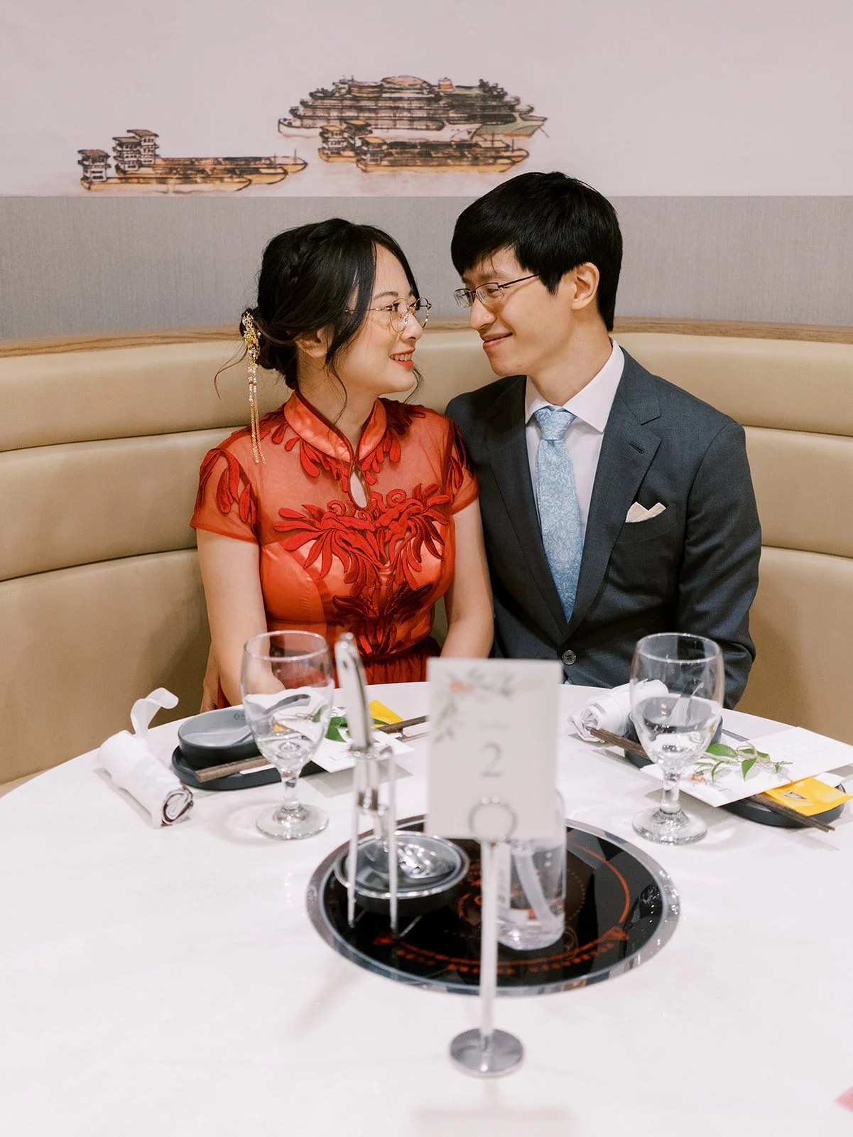 A bride and groom dressed in formal attire, sitting at a dinner table, gazing at each other, in an elegant restaurant setting.