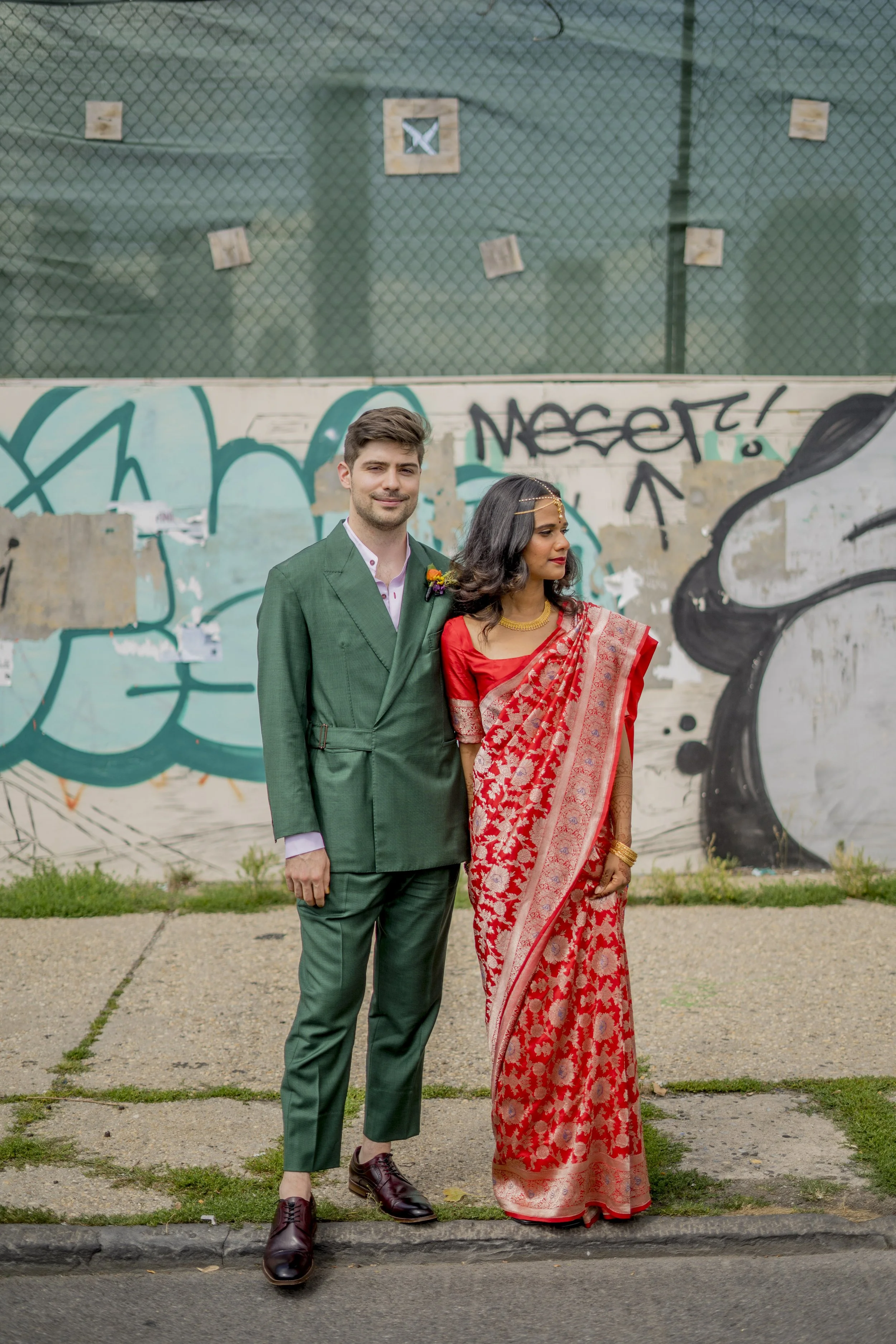 A wedding couple dressed in formal attire standing in front of a graffitied wall. The man is wearing a green suit with a boutonniere, and the woman is in a traditional red and gold saree with jewelry and makeup.