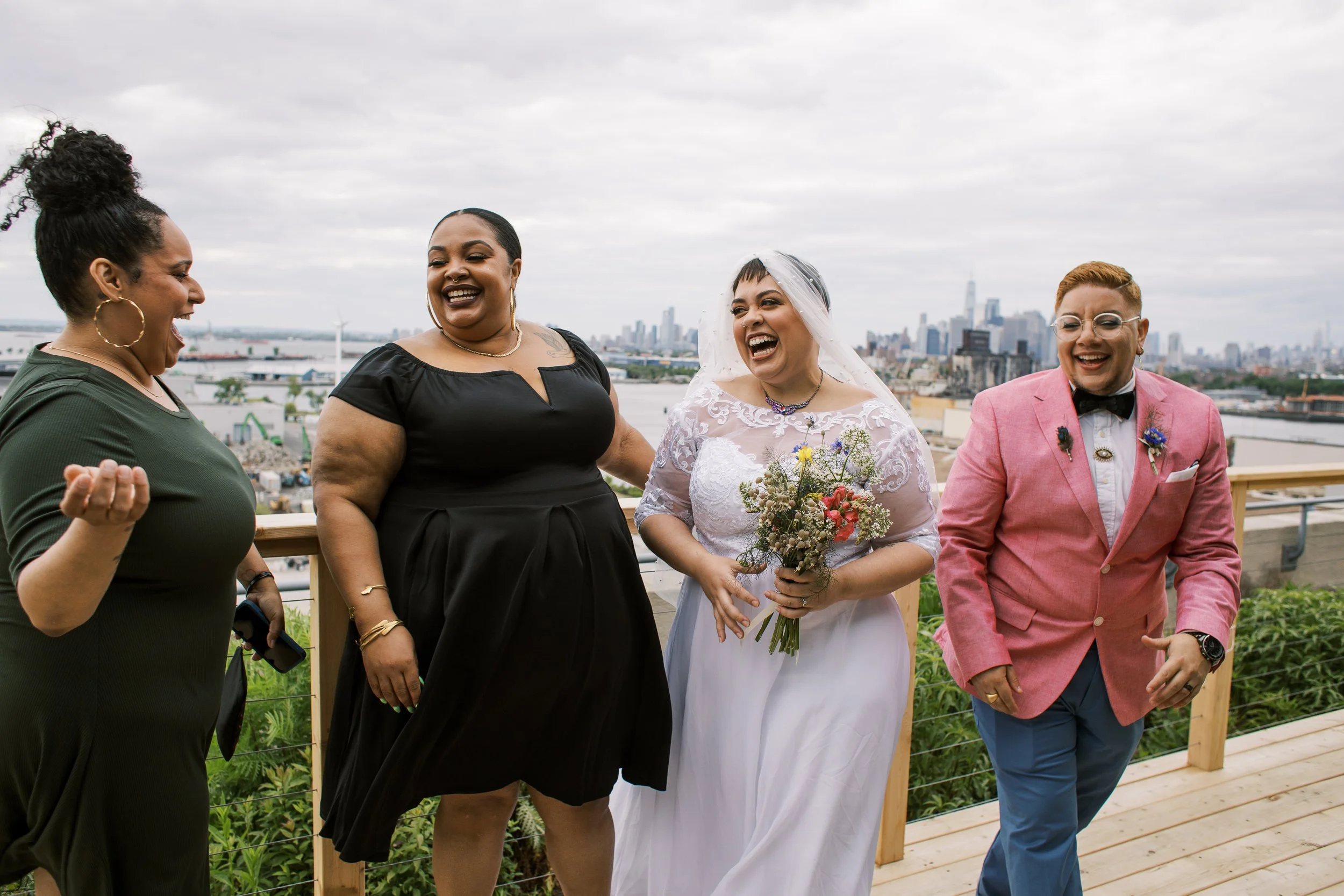 Group of five people, including a bride in a white wedding dress holding a bouquet, laughing and enjoying a celebration on a rooftop with city skyline in the background.