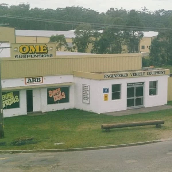 Exterior view of an automotive parts store with signs for OME Suspensions, ARB, and Engineered Vehicle Equipment, with a grassy lawn and a bench in front.