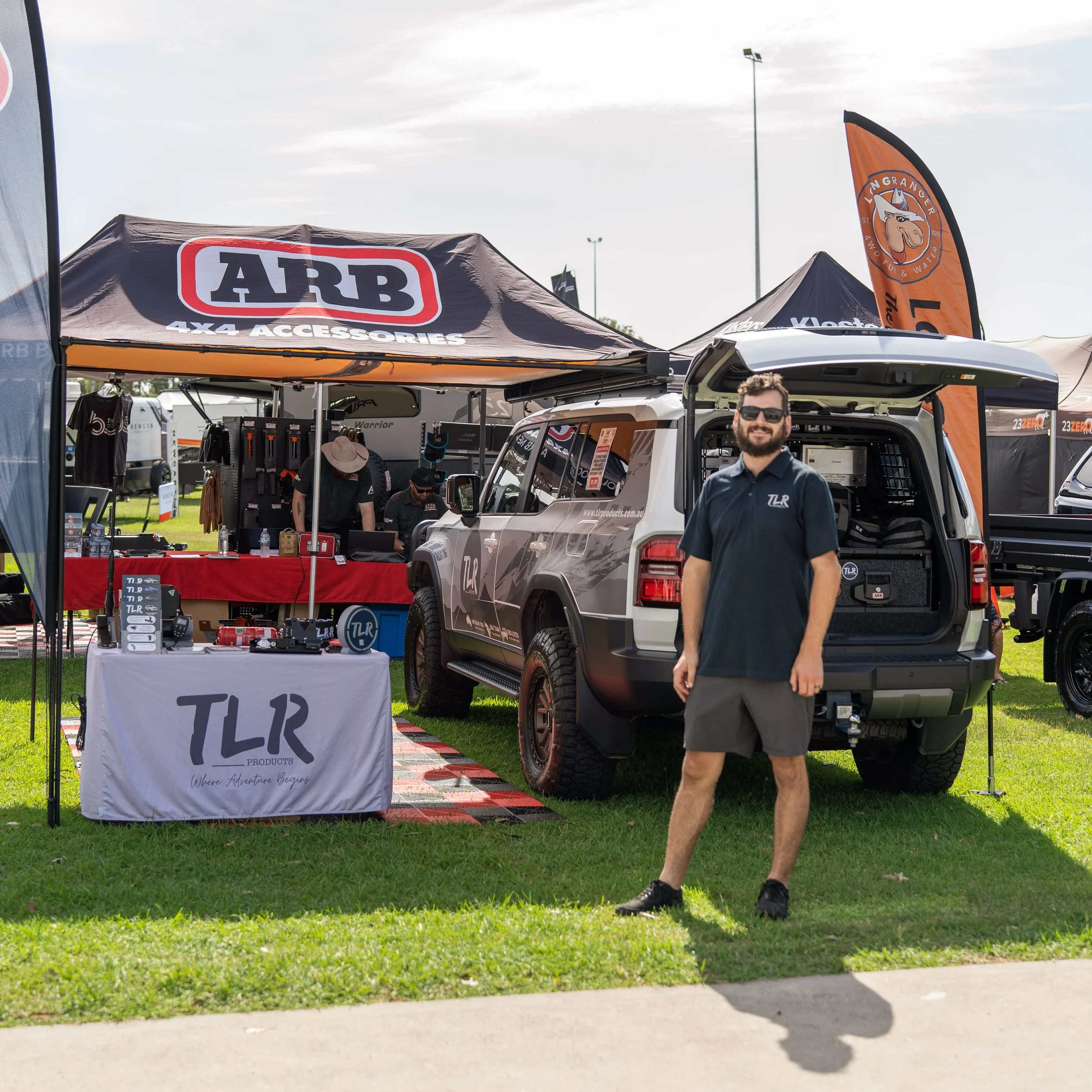 A man in sunglasses standing next to the open trunk of a vehicle at an outdoor event with tents, flags, and other people visible in the background.