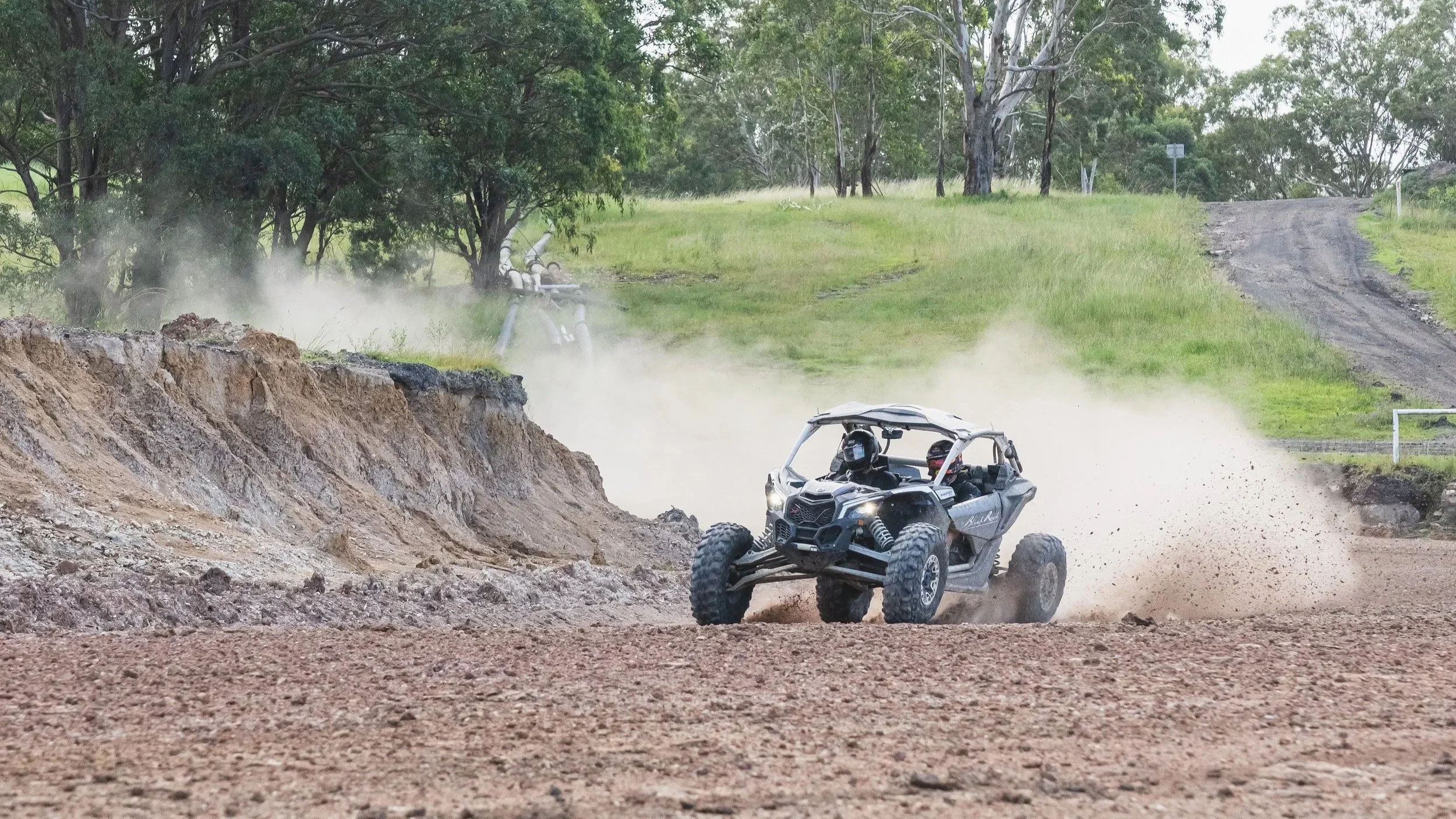An off-road utility vehicle driving on dirt trail in a wooded area, kicking up dust with trees and grassy fields in the background.