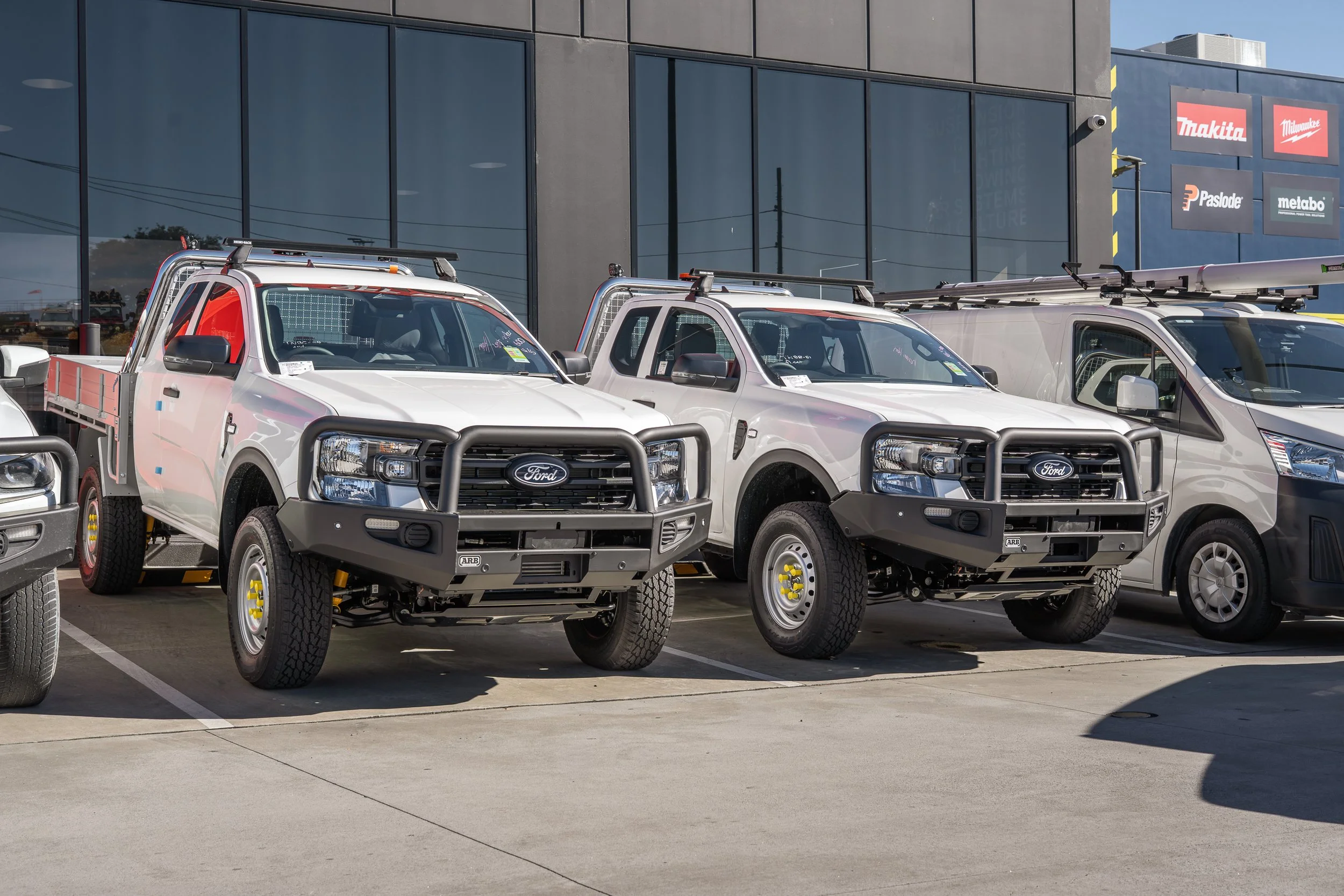 Three white pickup trucks parked outside a building with large glass windows and signage for brands like Makita, Paslode, and Metabo.