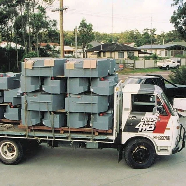 A flatbed truck carrying multiple gray concrete blocks parked on a residential street, with houses and trees in the background.