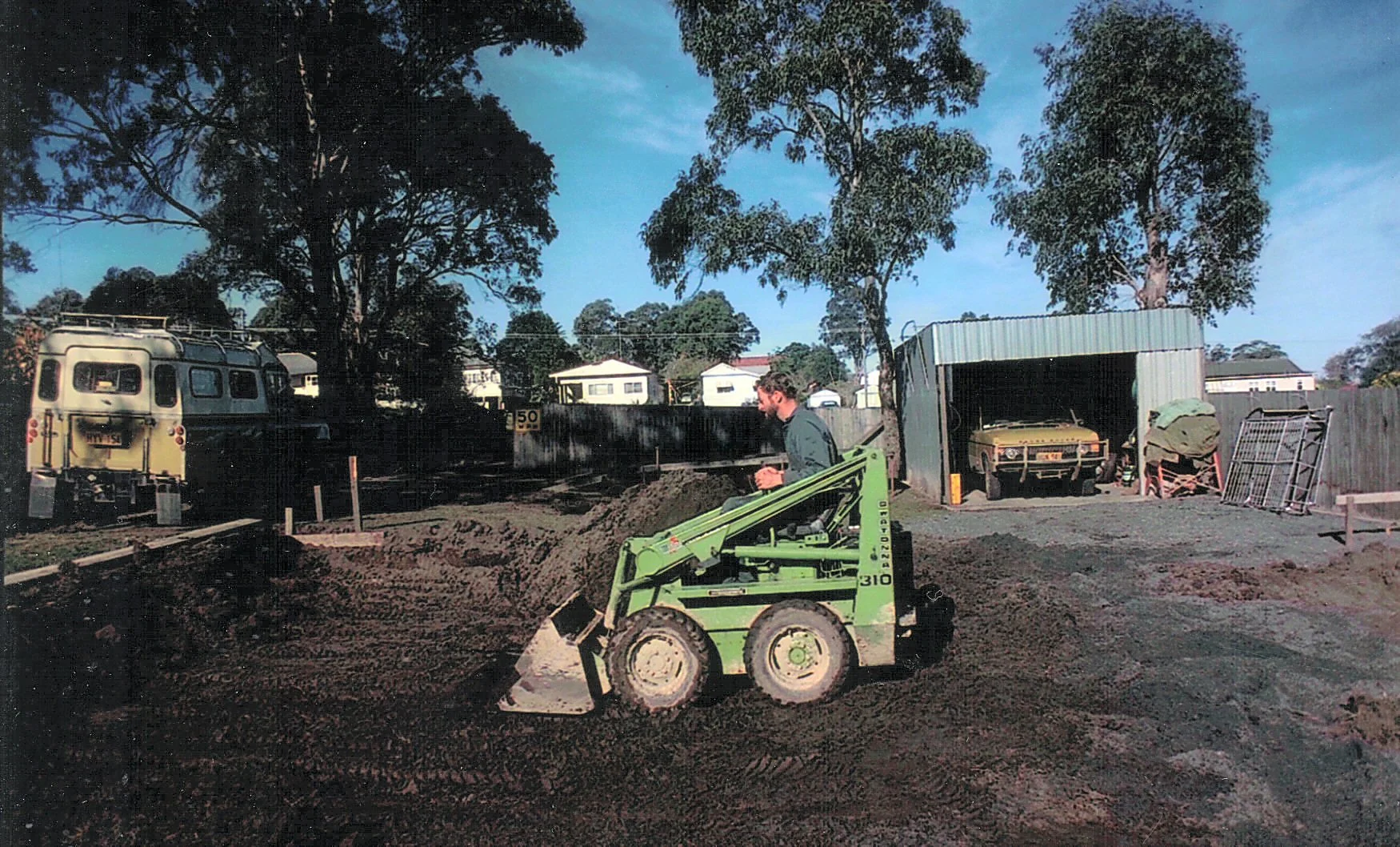 A man operating a small green construction vehicle on dirt ground in a backyard with trees, a fence, a shed, and two parked cars, one in the shed and one outside, during daytime.