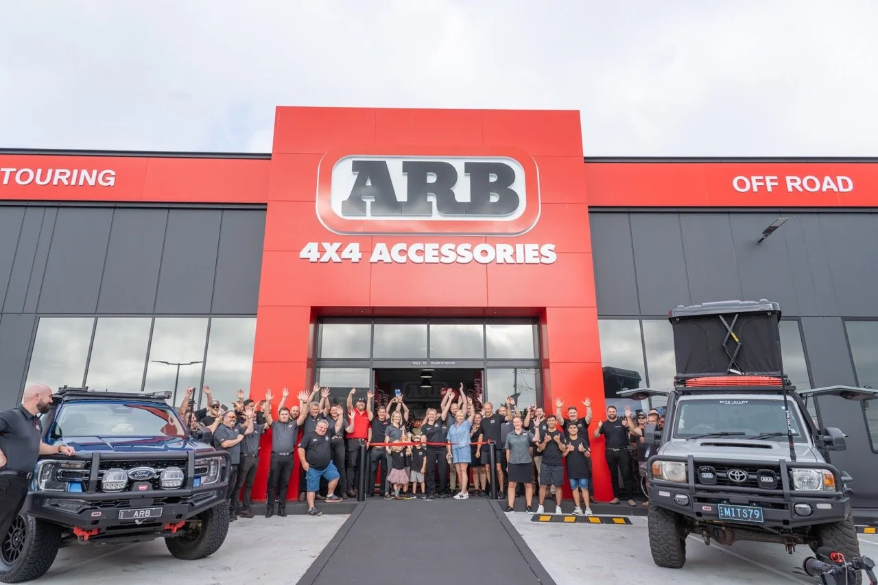 Grand opening of an ARB store with a group of employees and customers in front, two off-road vehicles parked outside, and a red and black building.