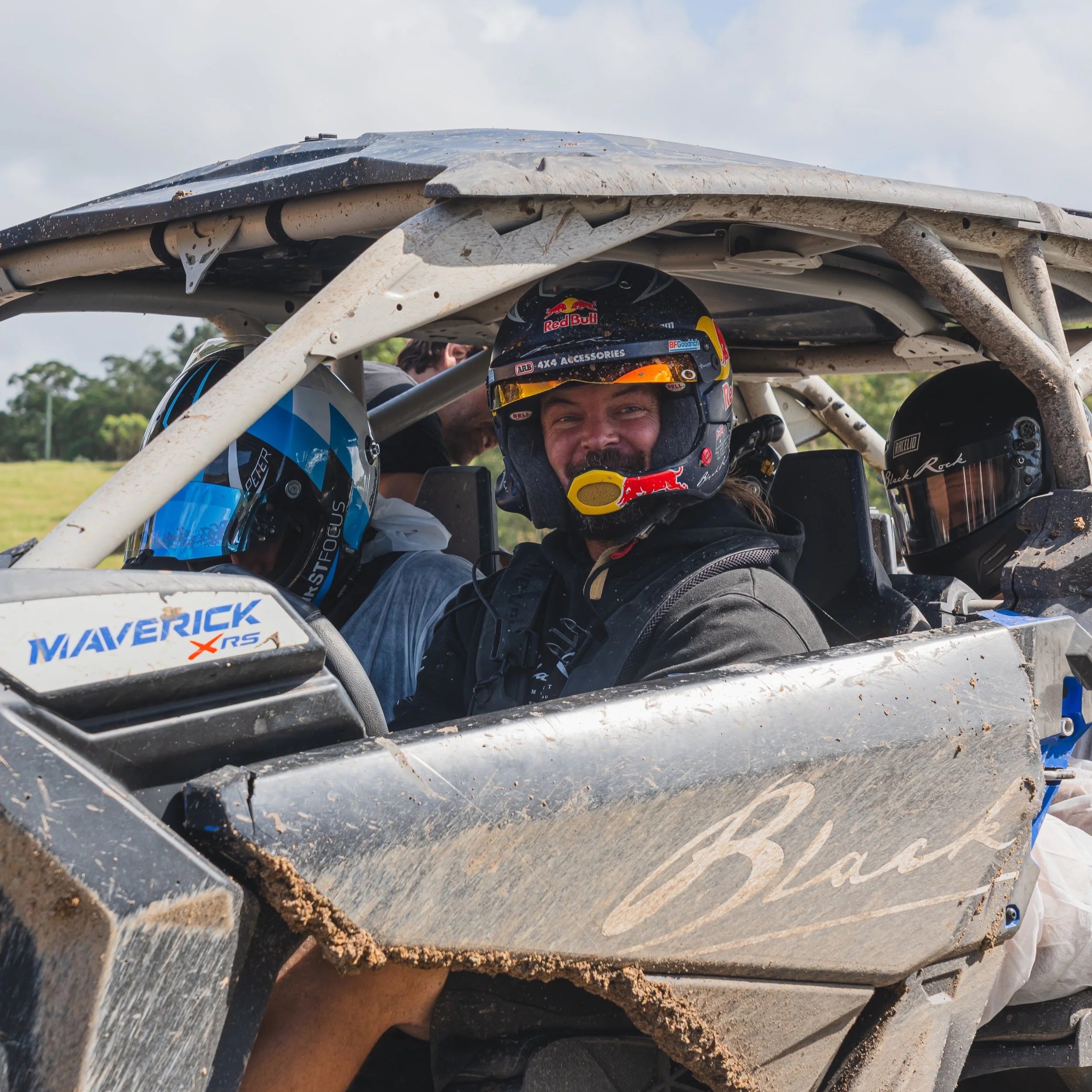 Close-up view of a dirt-covered off-road vehicle with a smiling man wearing a helmet inside. The vehicle has a white frame and mud-splattered decals, including "MAVERICK X RS."
