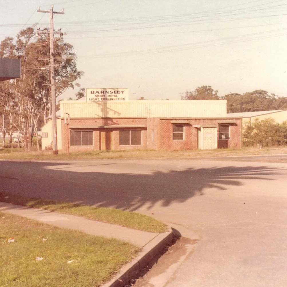 An old, small brick building with a sign that reads "Barnsley Sheet Metal Light Construction," situated on a corner with a sidewalk and grassy area. There are trees and utility poles with wires in the background.