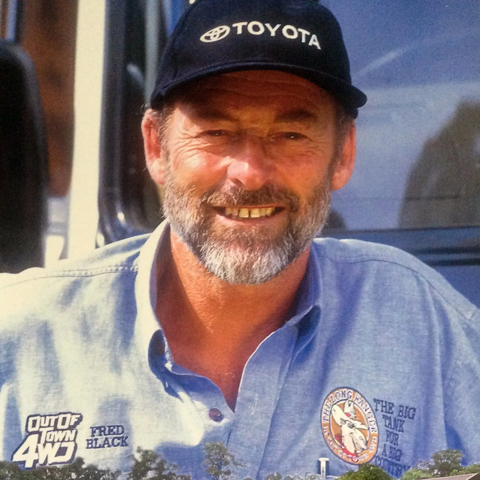 A smiling man with a beard wearing a blue Polo shirt and a black cap with the Toyota logo. The shirt has various logos and patches on it.