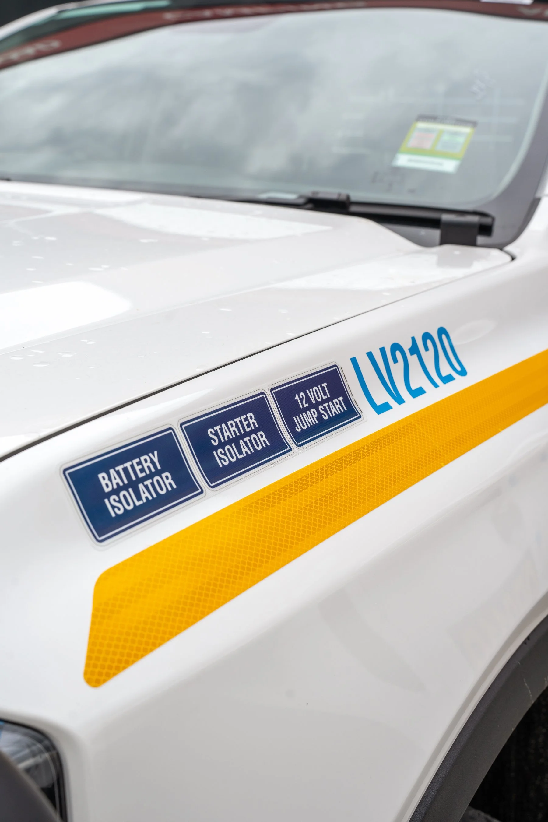 Close-up of a white vehicle with blue and yellow warning labels that read 'Battery Isolator' and '12 Volt Jump Start'.