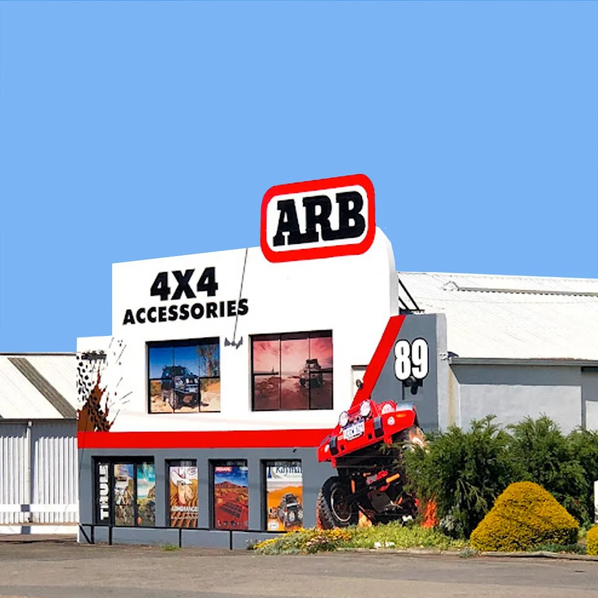 A building with signs that say 'ARB,' '4X4 ACCESSORIES,' and the number '89,' featuring off-road vehicle images, a motorcycle, and outdoor decorations, with a clear blue sky background.