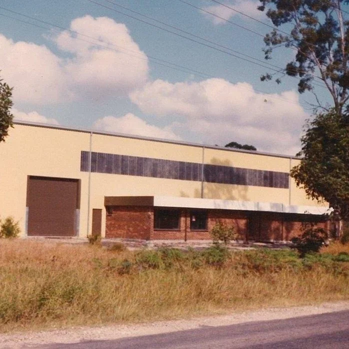 A large, beige industrial building with a brown section on the front, featuring a rolled door, brick wall, and windows. There is grassy land and trees in front, and power lines in the sky.