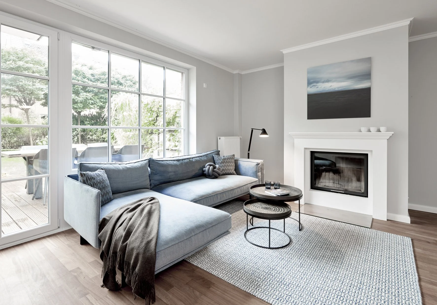 Wohnzimmer Kamin weiss Fotokunst blau Teppich miinu Sofa blau Sofatische rund Ethnicraft Fensterfront weiss