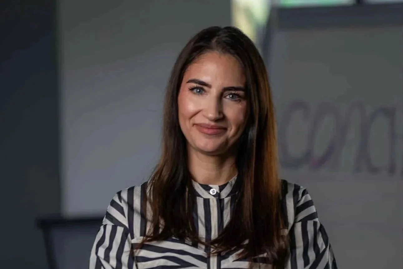 A woman with long dark hair, wearing a black and white striped top, smiling at the camera in an indoor setting, with a blurred background featuring a window and a whiteboard.