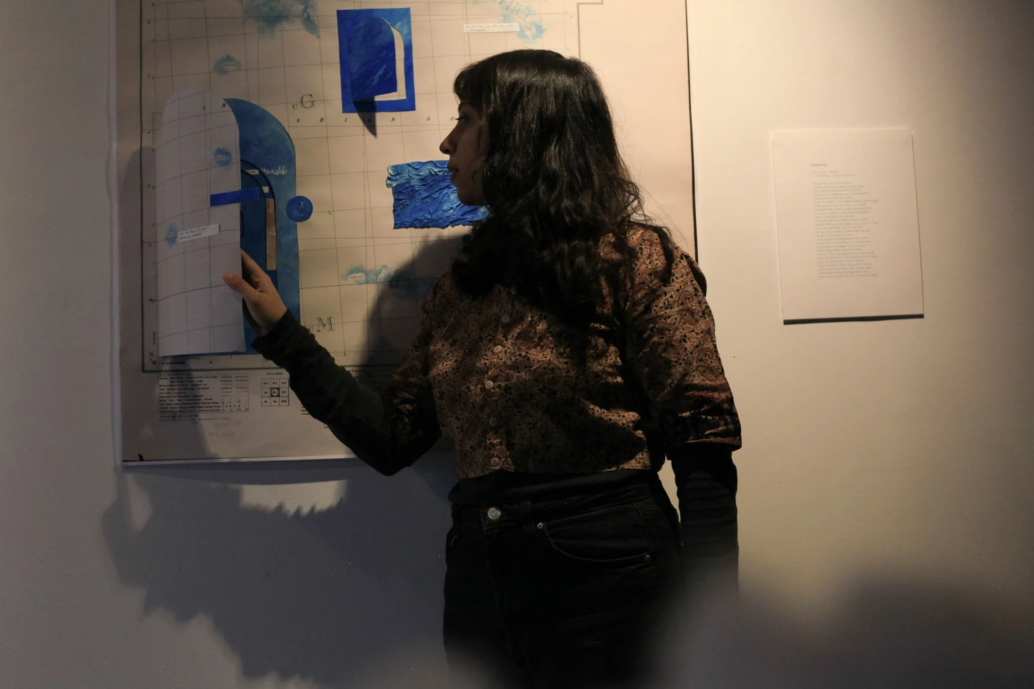 A brown femme person with medium length curly black hair holds open a door cut out from a large map they're standing in front of. There's hints of ocean blue all over the map. To their right hangs a poem.