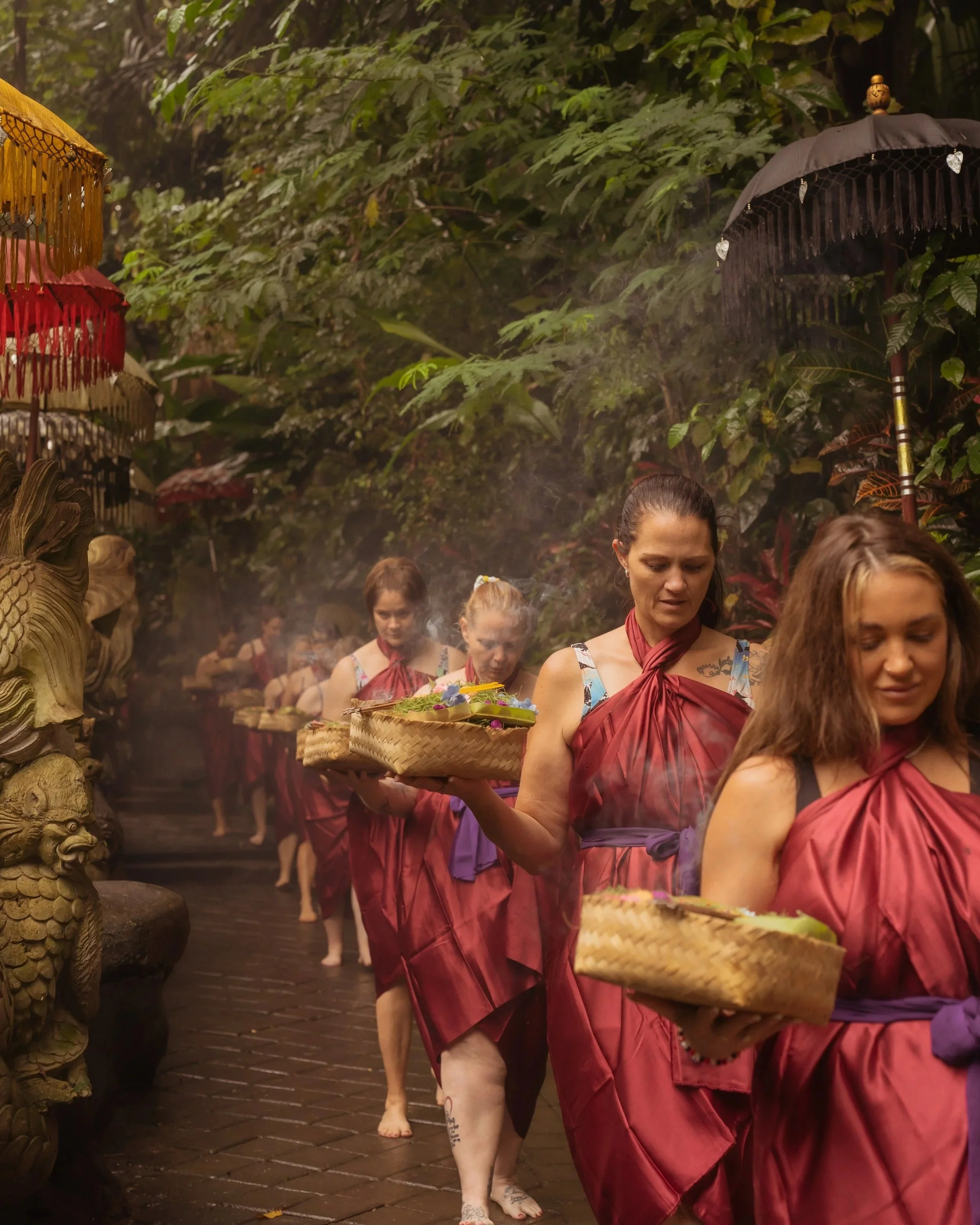Women in traditional robes holding baskets and umbrellas, walking through a lush, green tropical forest pathway during a ceremonial procession.