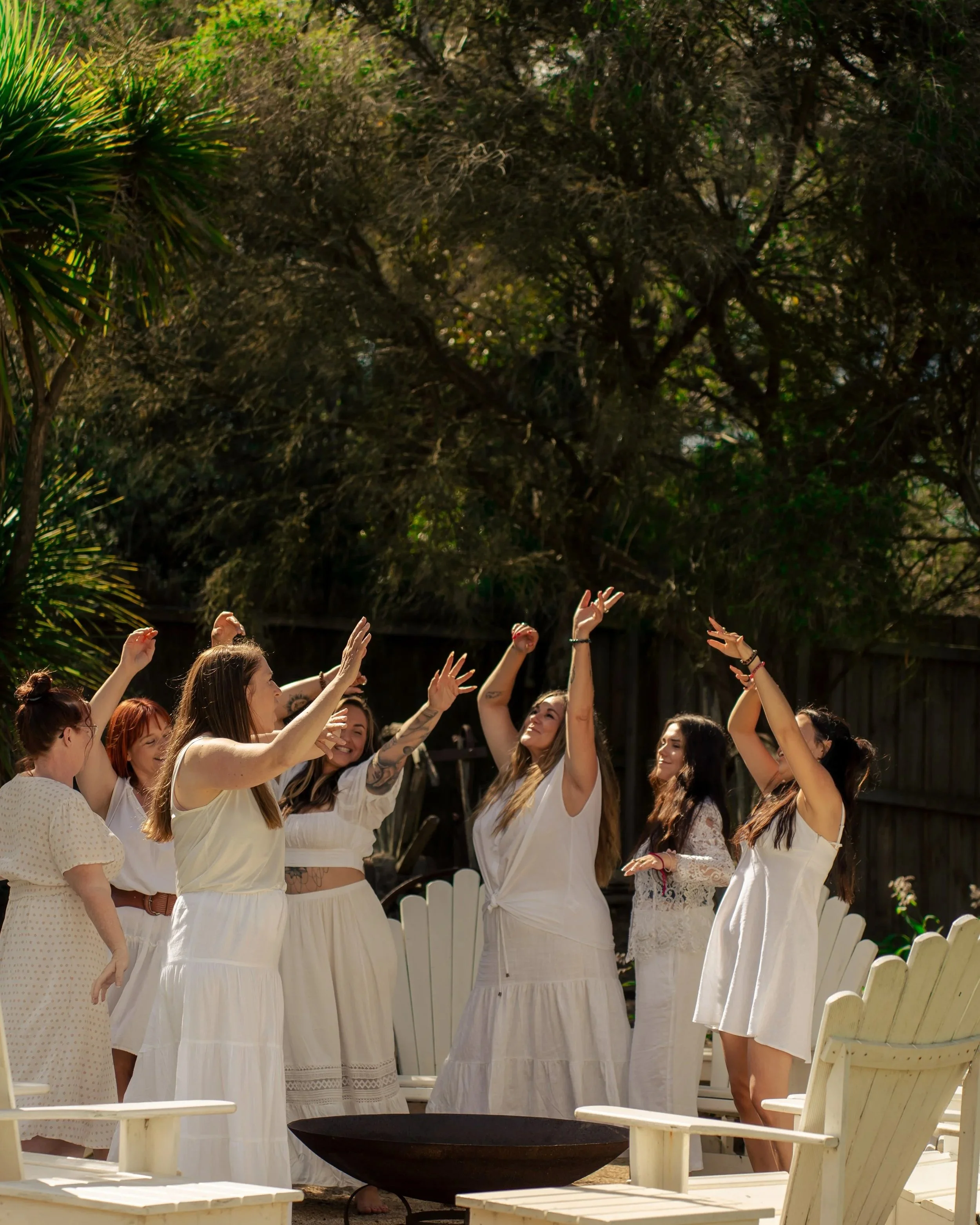 Women dancing and celebrating outdoors during daytime, surrounded by greenery and white Adirondack chairs.