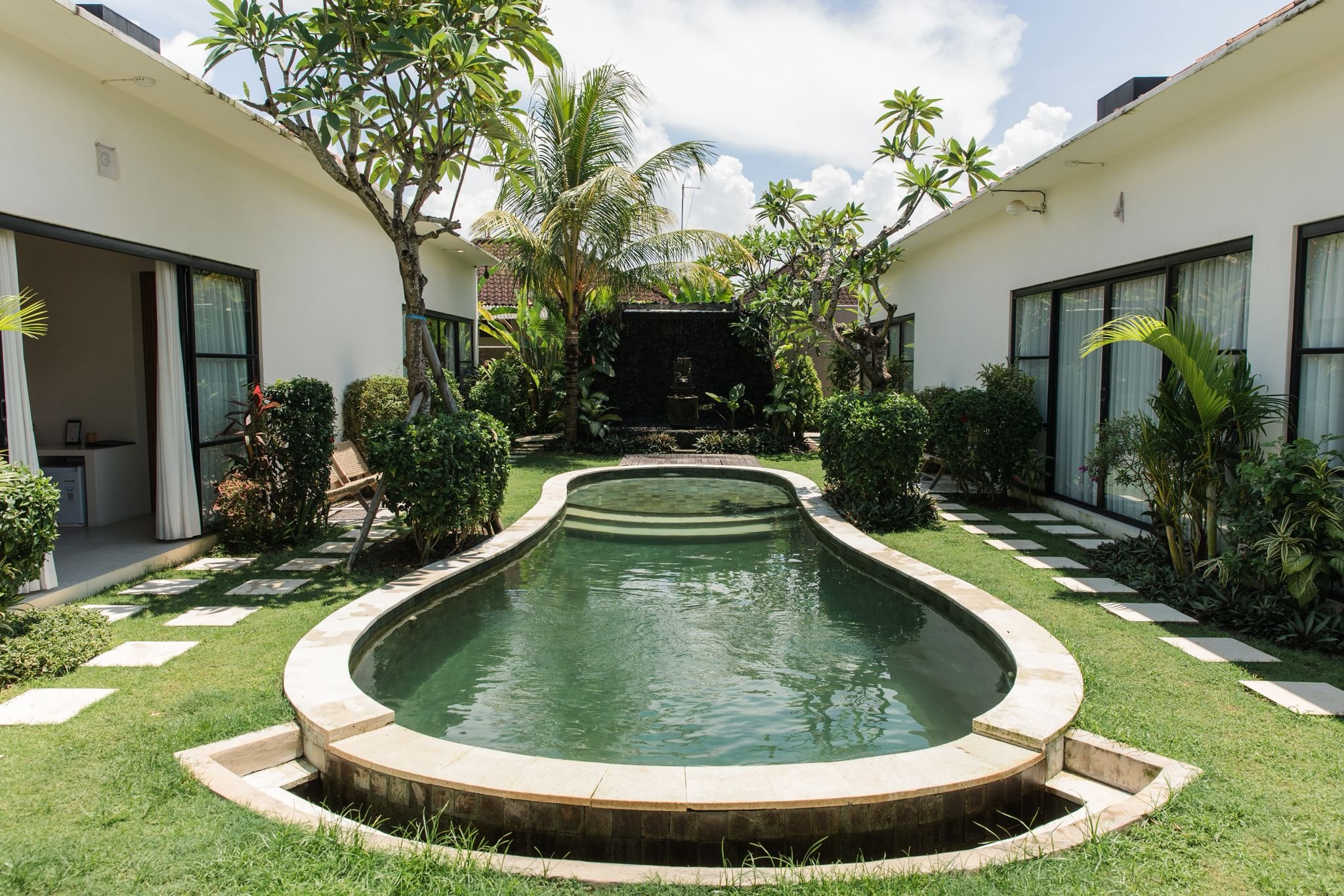 A narrow swimming pool in a backyard courtyard surrounded by greenery and white buildings with sliding glass doors, with trees and plants, under a partly cloudy sky.