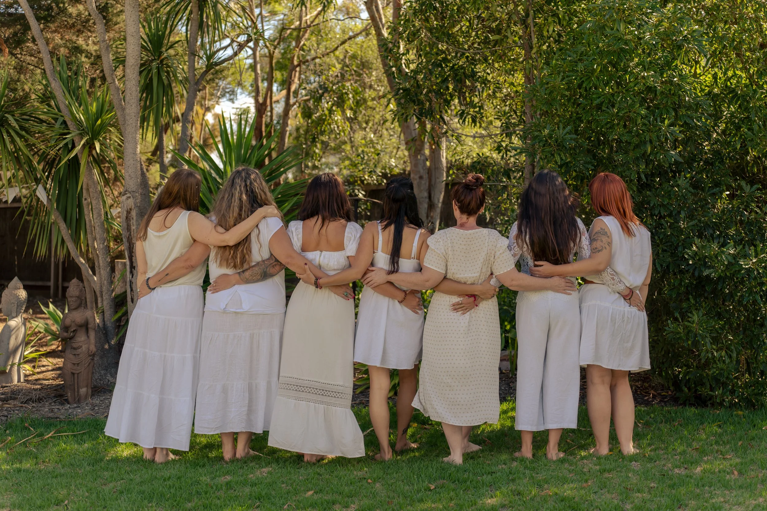 A group of women standing outdoors with their backs to the camera, embracing each other, surrounded by trees and greenery.