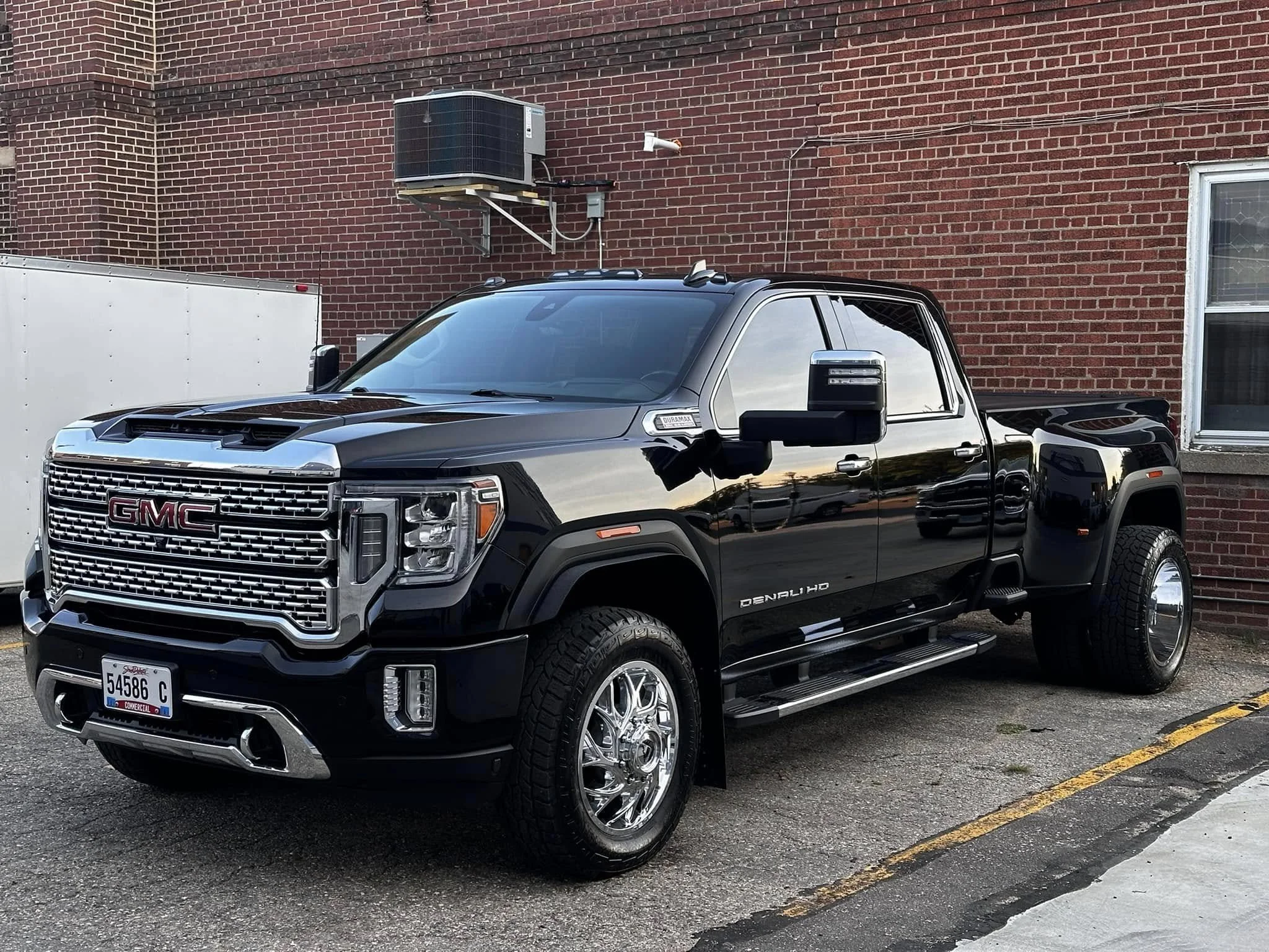 A black GMC Sierra Denali HD pickup truck parked beside a brick building, with a white trailer on the left and an air conditioning unit above it.