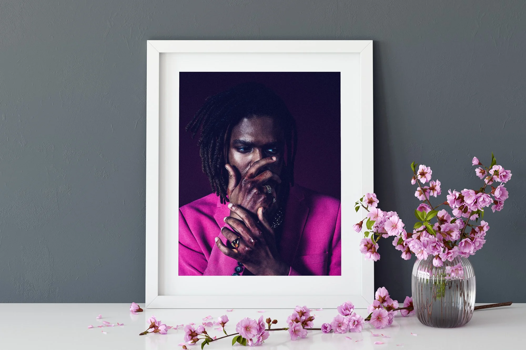 A framed portrait of a man with dreadlocks wearing a pink blazer, surrounded by pink flowers in a glass vase and scattered on a white surface.