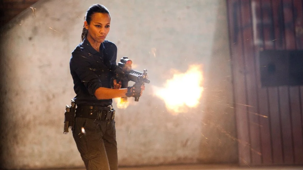 A woman dressed in black tactical clothing firing a gun with a gunshot creating a flash and sparks inside a warehouse or industrial setting.