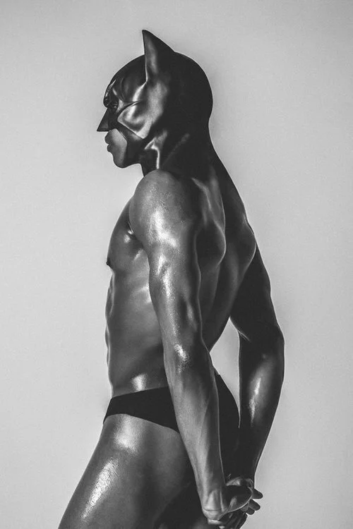 A muscular man wearing a Batman mask, shirtless, in a side profile pose with glossy skin, black underwear, and his hands behind his back against a plain background.