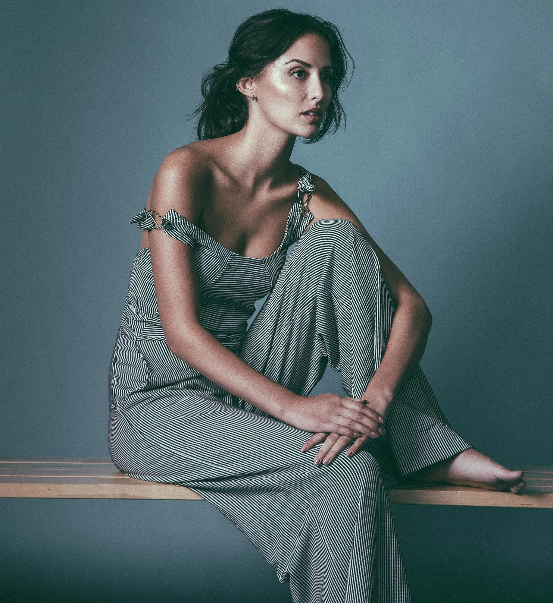 A woman with dark hair and fair skin wearing striped pajamas sitting on a wooden bench against a plain blue-gray background.