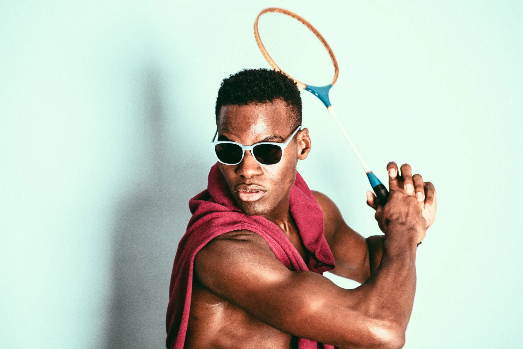 A young man in sunglasses holding a badminton racket over his shoulder, wearing a sleeveless hoodie, against a light background.