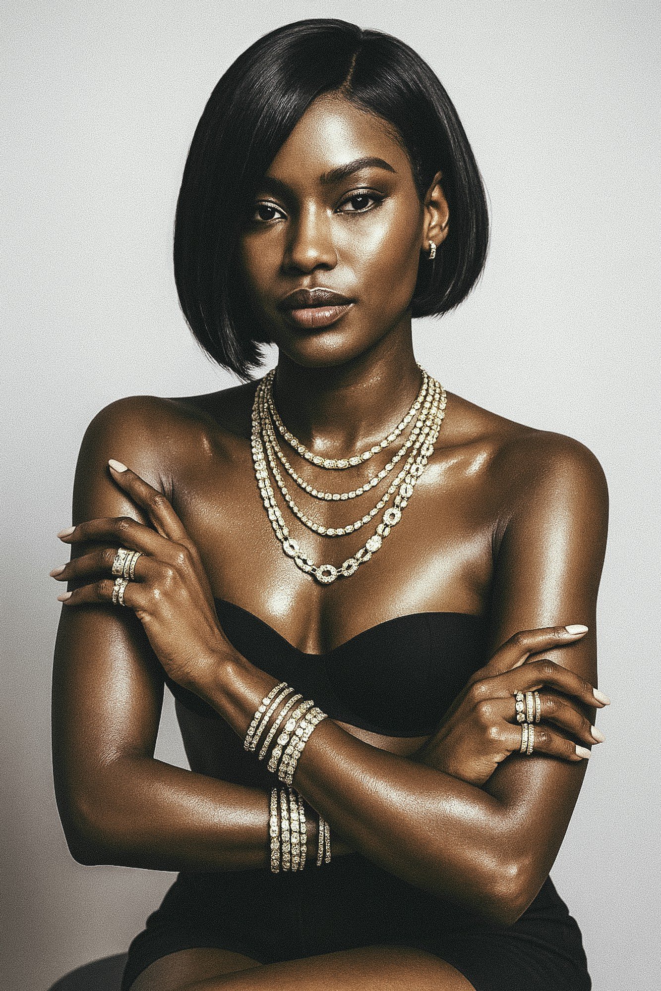 A woman with dark skin and short black hair styled in a bob, wearing multiple pearl necklaces, matching rings and bracelets, and a black strapless top against a light gray background.
