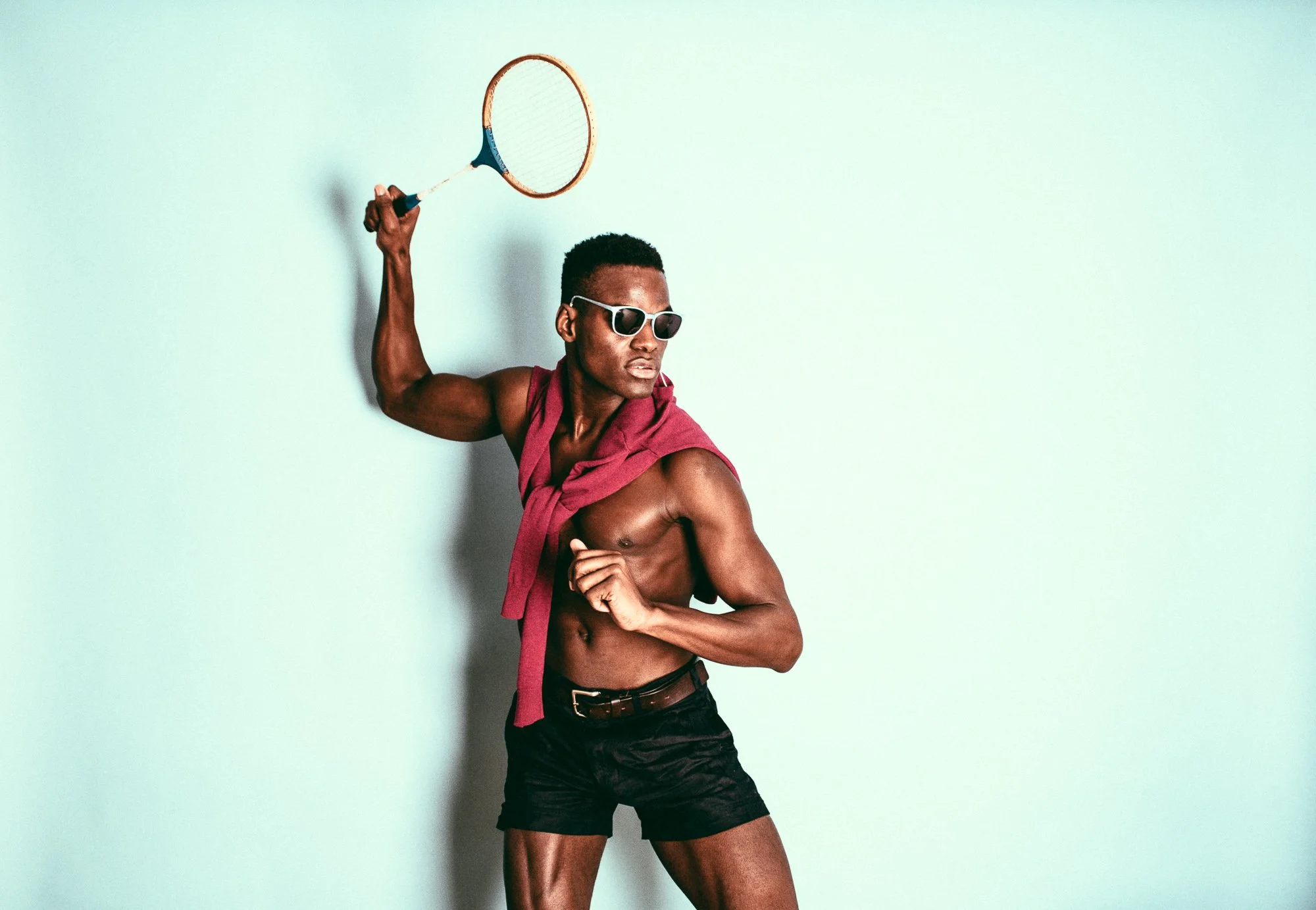 A muscular man wearing sunglasses, black shorts, and a red shirt draped over his shoulders, holding a tennis racket in a dynamic pose against a plain backdrop.