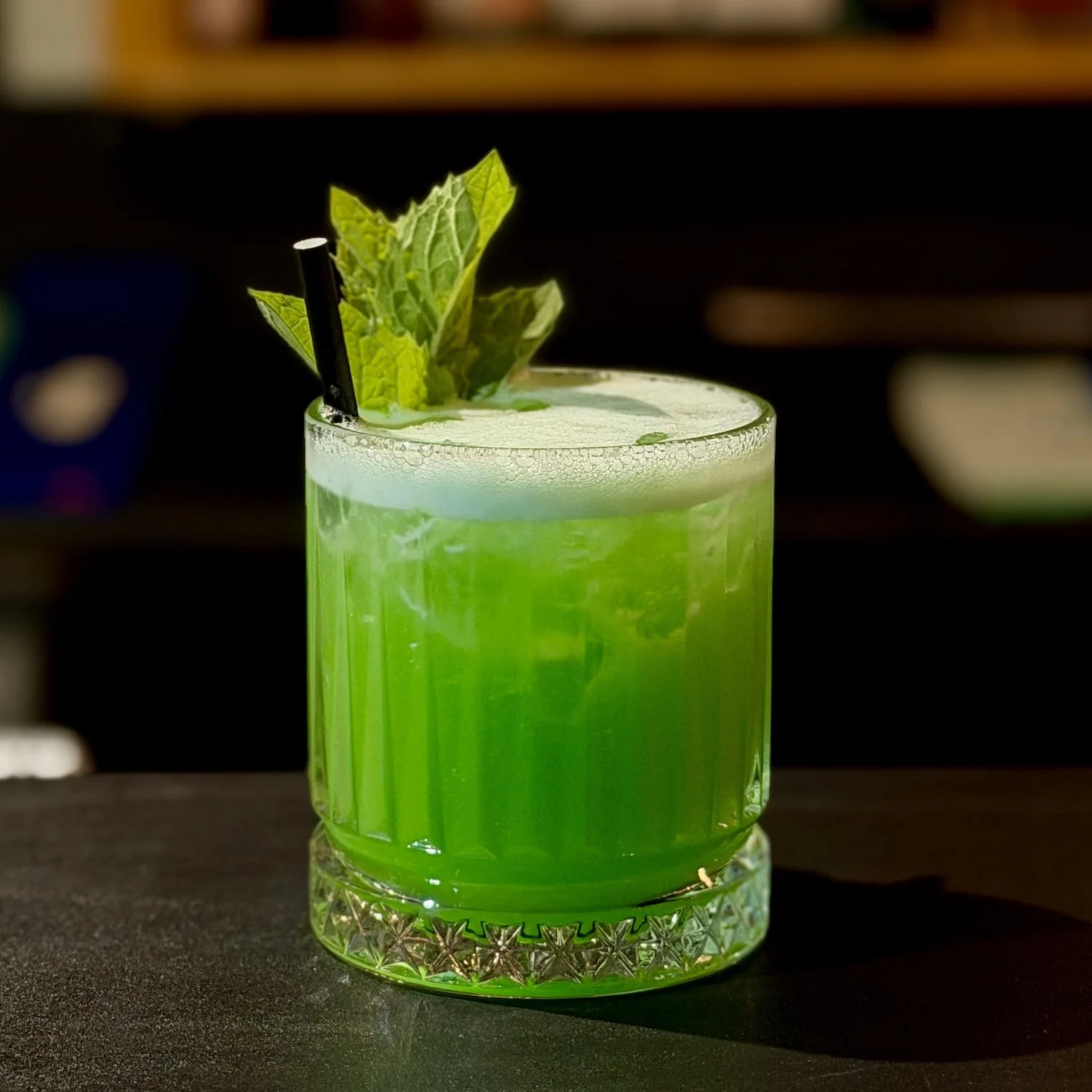 A vibrant green cocktail garnished with mint leaves and a black straw, served in a textured glass.