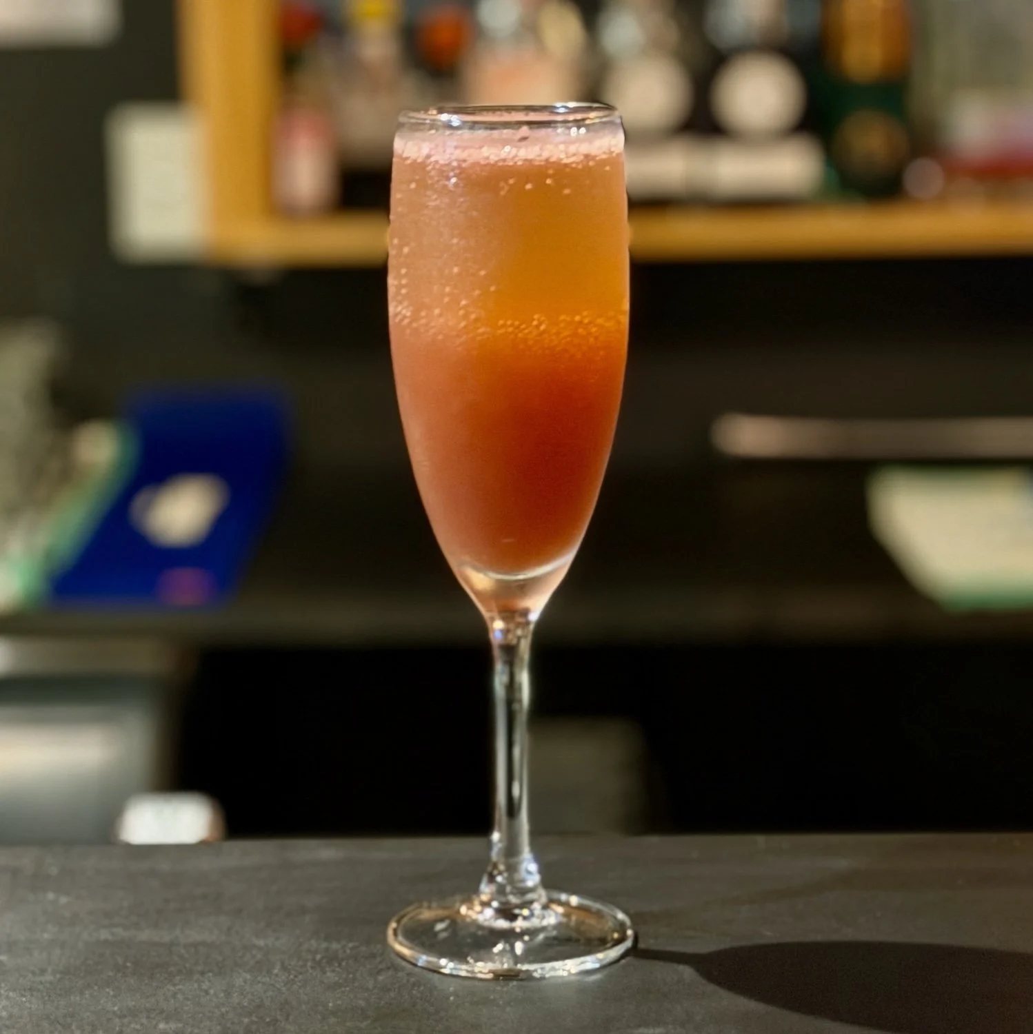 A tall champagne flute filled with a pinkish cocktail, with condensation on the outside, placed on a dark surface in a bar setting.