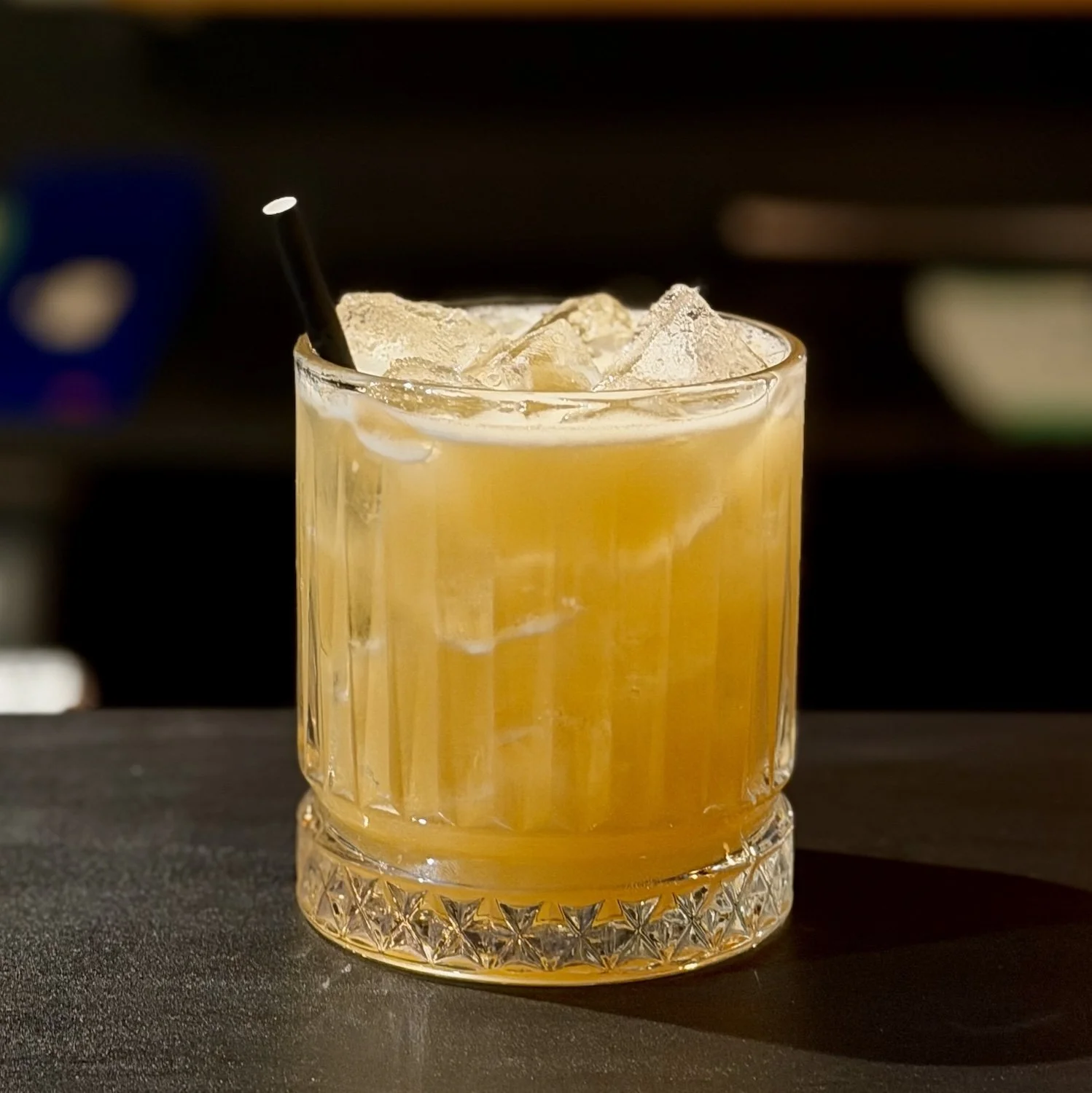 A glass with a yellow-orange cocktail and ice cubes, with a black straw, on a dark surface.