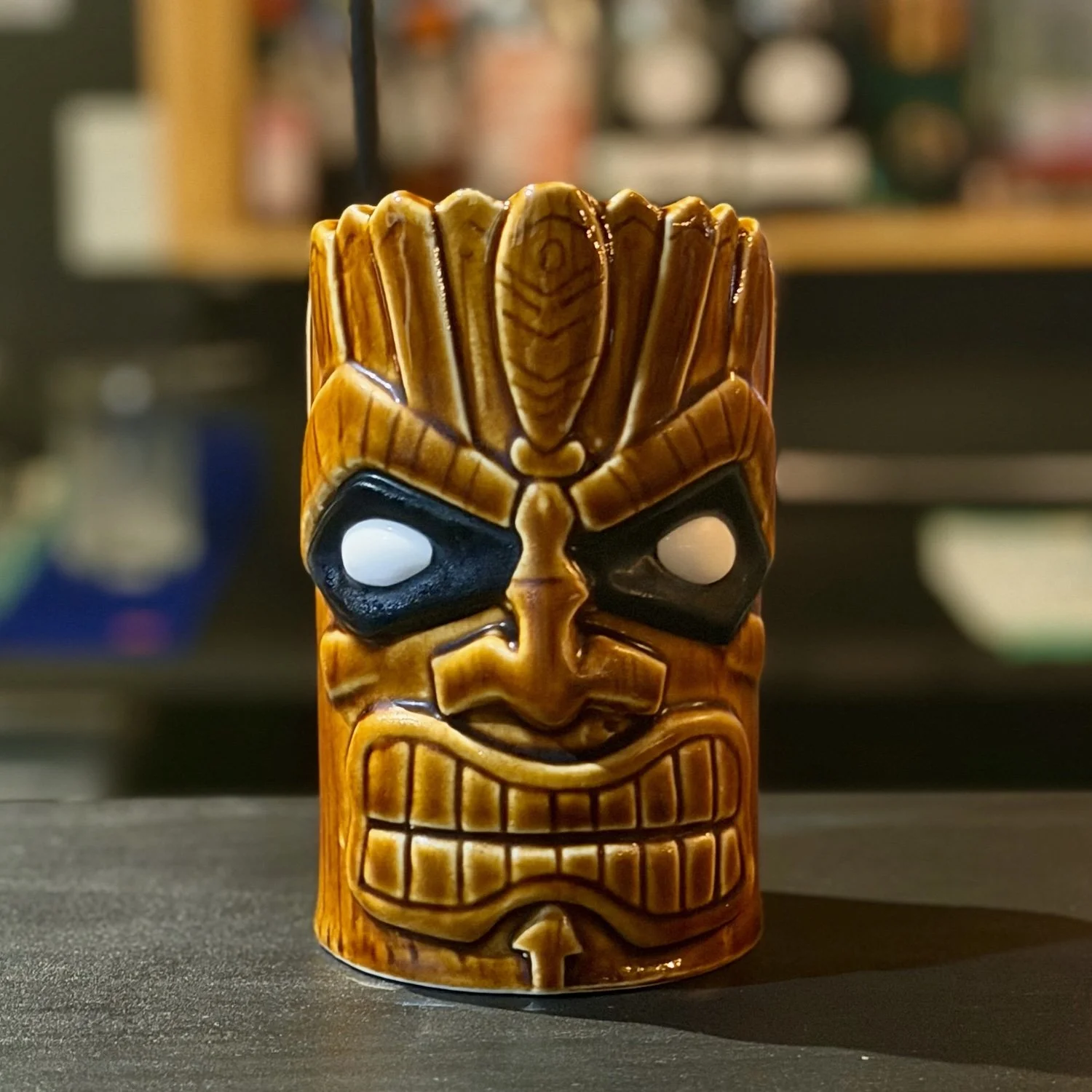 A tiki mug with a carved face resembling a tribal warrior, featuring exaggerated facial features and painted details, placed on a dark surface.