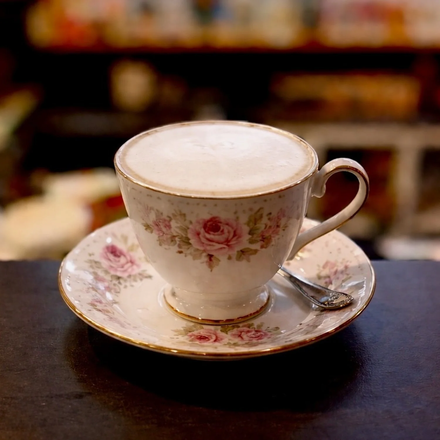 A floral-patterned teacup filled with a frothy beverage, placed on a matching saucer with a small spoon on a dark surface.