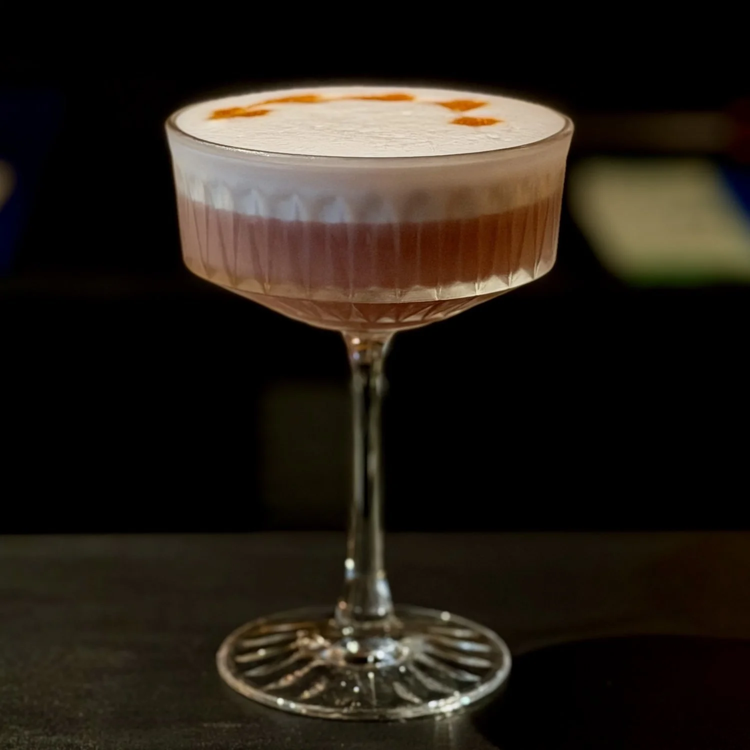 A cocktail in a coupe glass with white foam topped with orange peel twist garnishing the drink.
