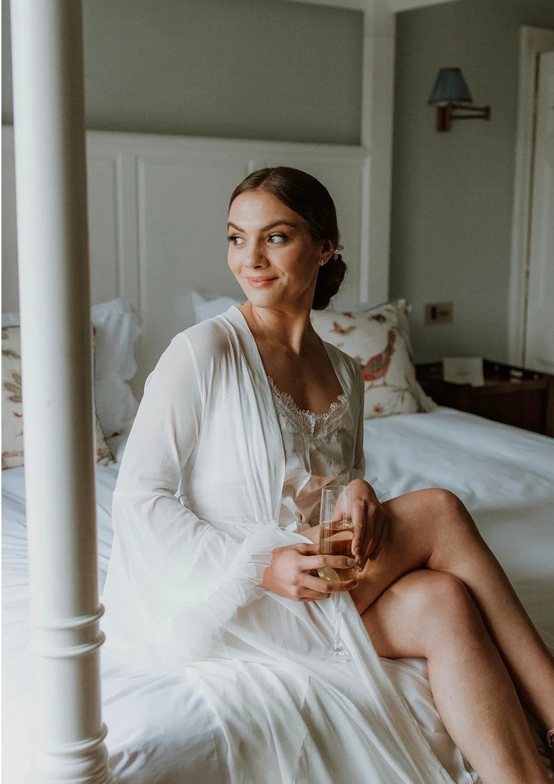 Bride sitting on a bed with a glass of champagne in hand with her natural makeup look complete and hair in a low bun