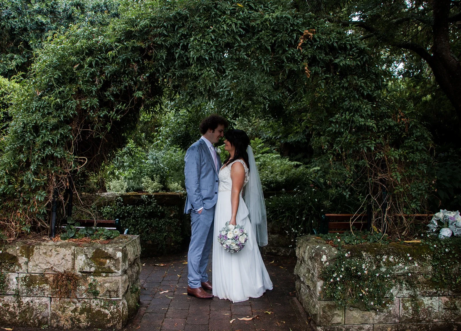 A bride and groom standing close together in a wooded outdoor setting, holding hands, with their foreheads touching, during their wedding photoshoot.