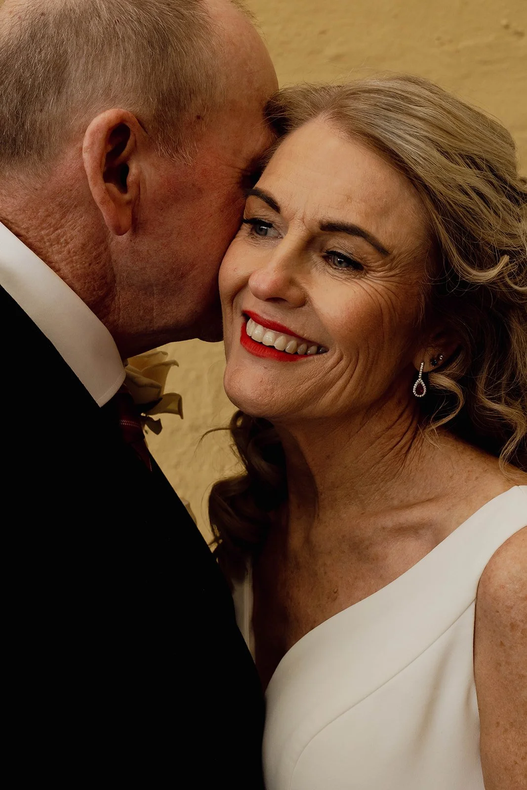 A man in a tuxedo with a boutonnière leans in to kiss a smiling woman in a white dress with jewelry, against a beige background.