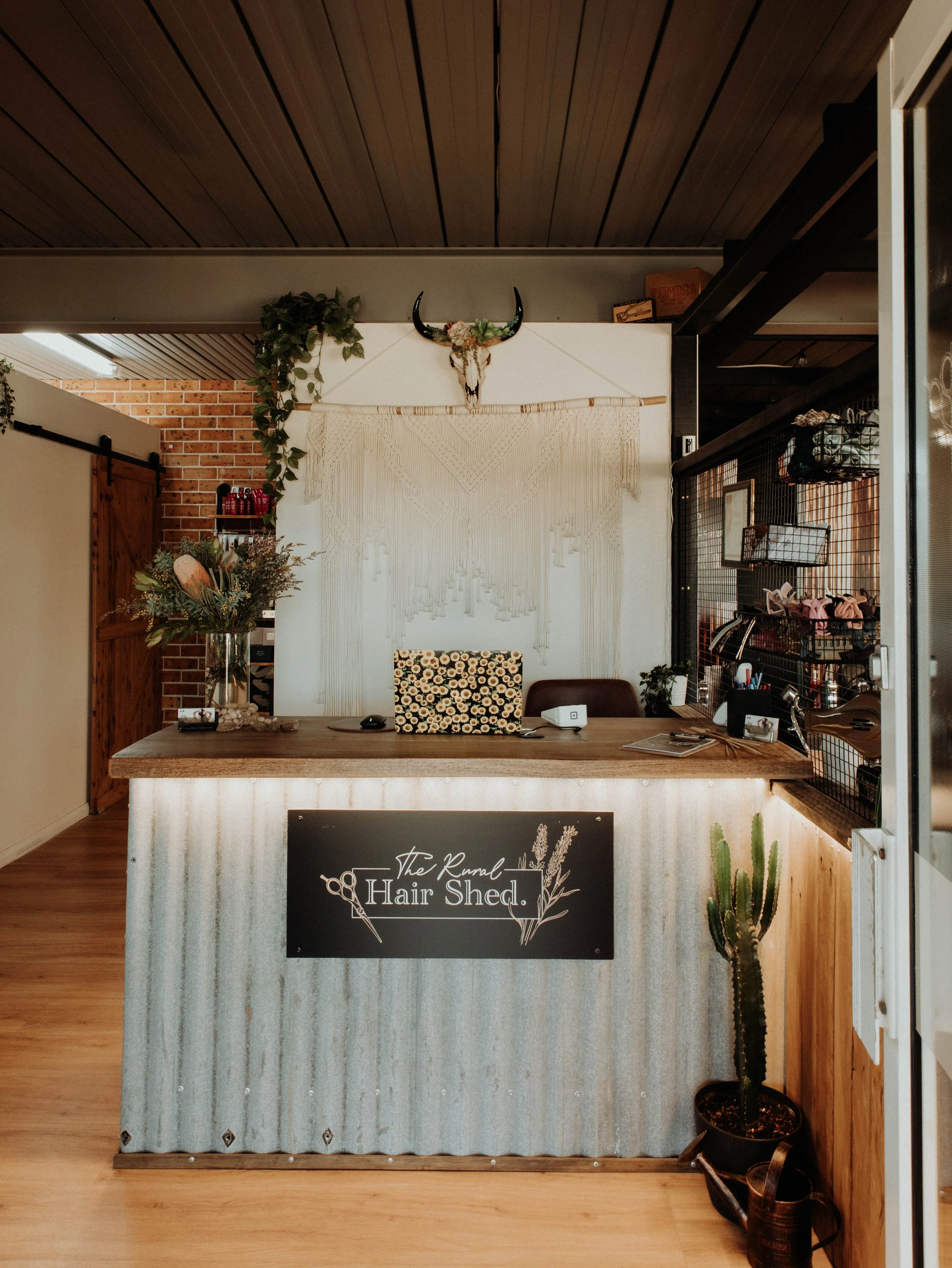 Reception desk at The Rural Hair Shed with a cactus plant on the right, a bouquet of flowers on the left, a decorative skull with horns and greenery on the wall behind, and a macrame wall hanging behind the desk.