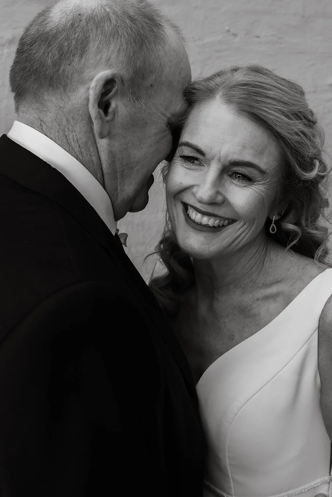A black and white photo of an older man in a tuxedo kissing a smiling woman in a white dress, likely at a wedding or formal event.