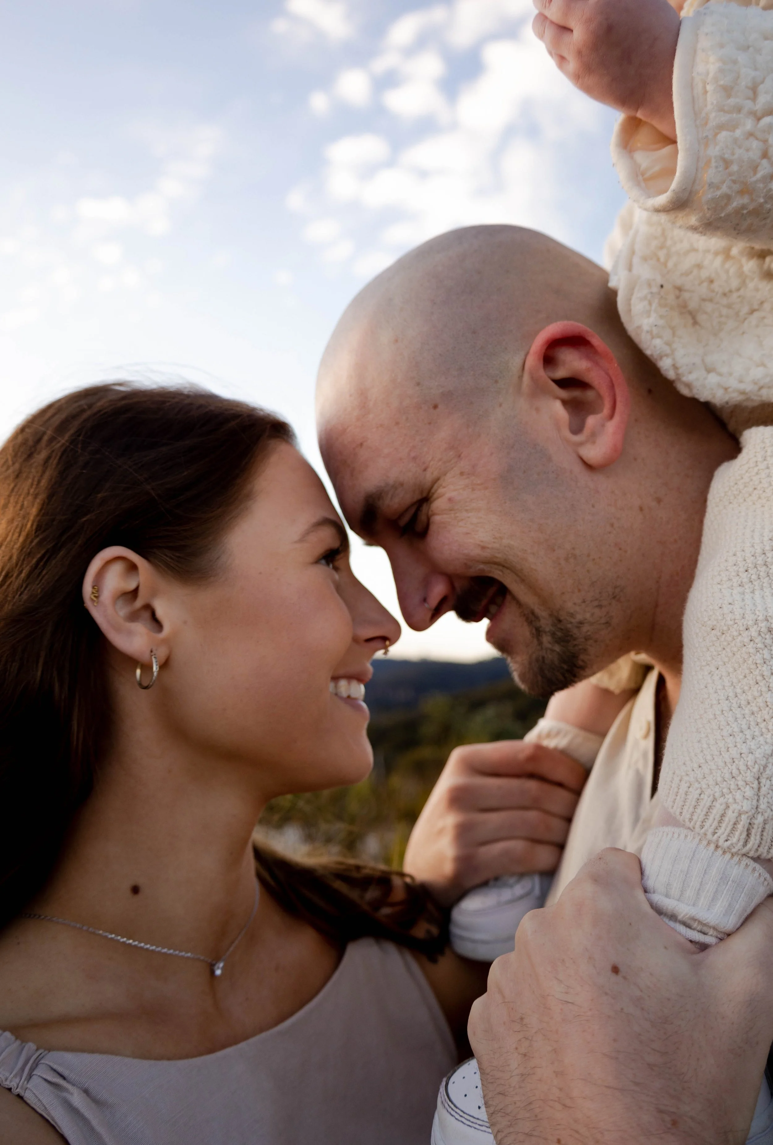 A woman and a man face each other with their foreheads touching, smiling and looking into each other's eyes, with a child on the man's shoulders. It is outdoors during sunset or late afternoon.