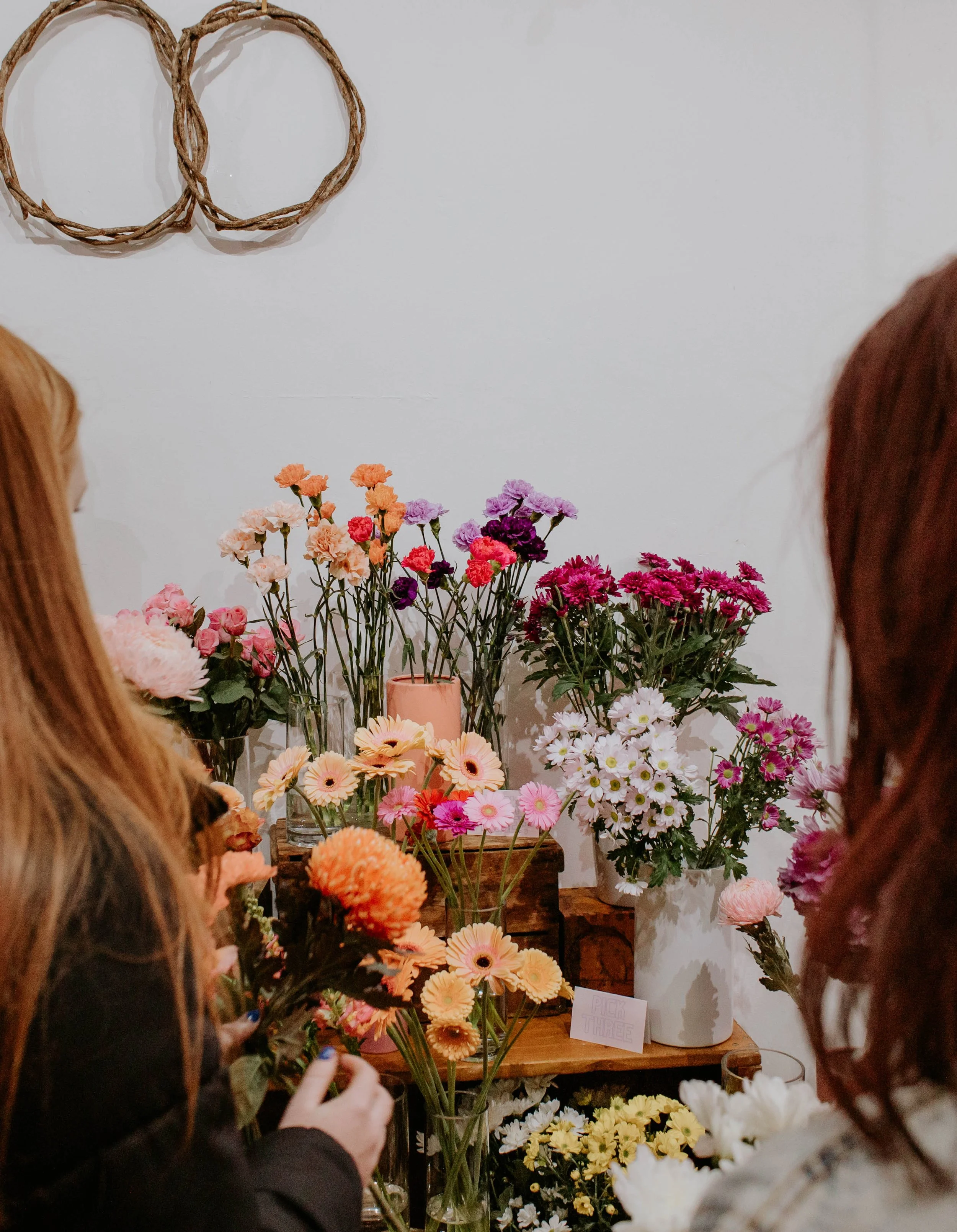 People browsing a display of colorful flowers at a flower shop or market. Flowers include daisies, carnations, and chrysanthemums in vases.