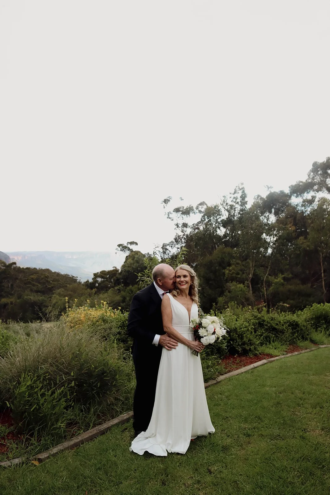 Bride and groom on their wedding day outdoors in a garden, with the groom kissing the bride on her cheek, she is holding a bouquet of white and red flowers, both smiling, greenery and trees in the background.