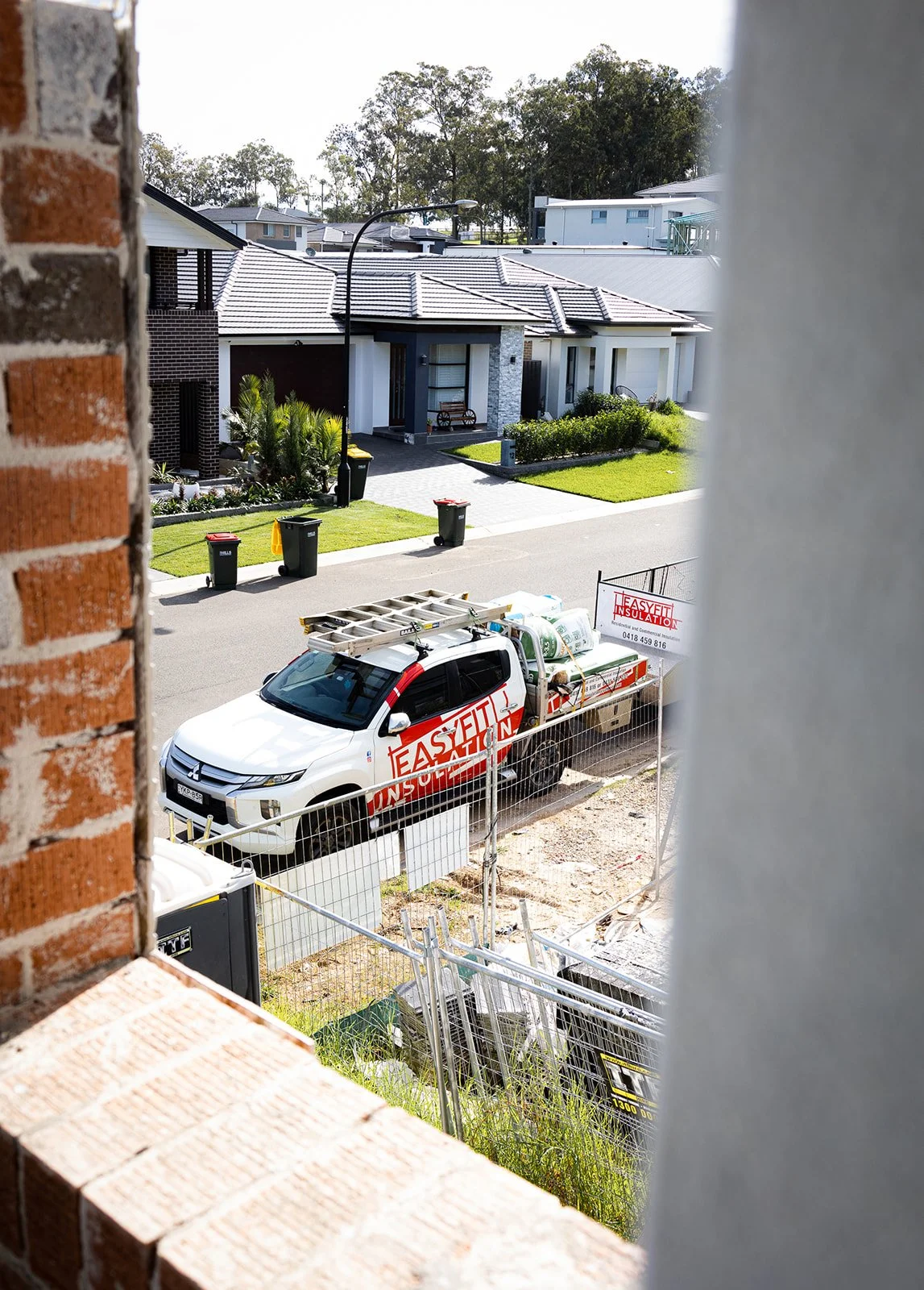 View from a window showing a white truck with red and black Onsite Fitting signage parked on a residential street with modern houses, green lawns, and trash bins.